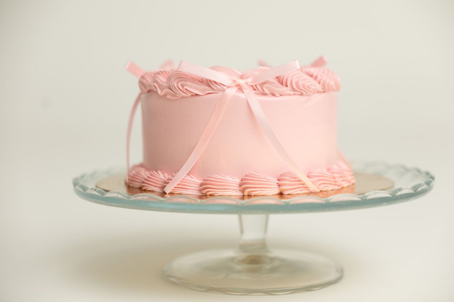 Pink frosted layer cake with ribbon bow on glass cake stand.