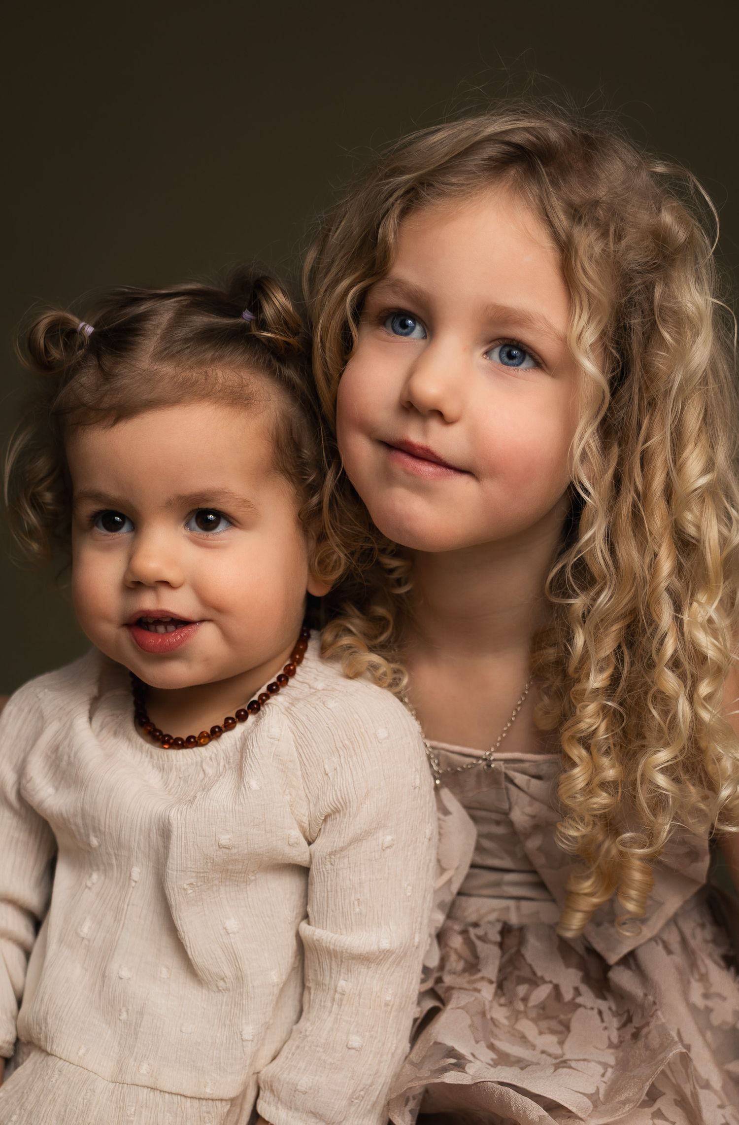 Two young girls posing together, older girl with curly blonde hair and blue eyes, younger toddler in cream dress.
