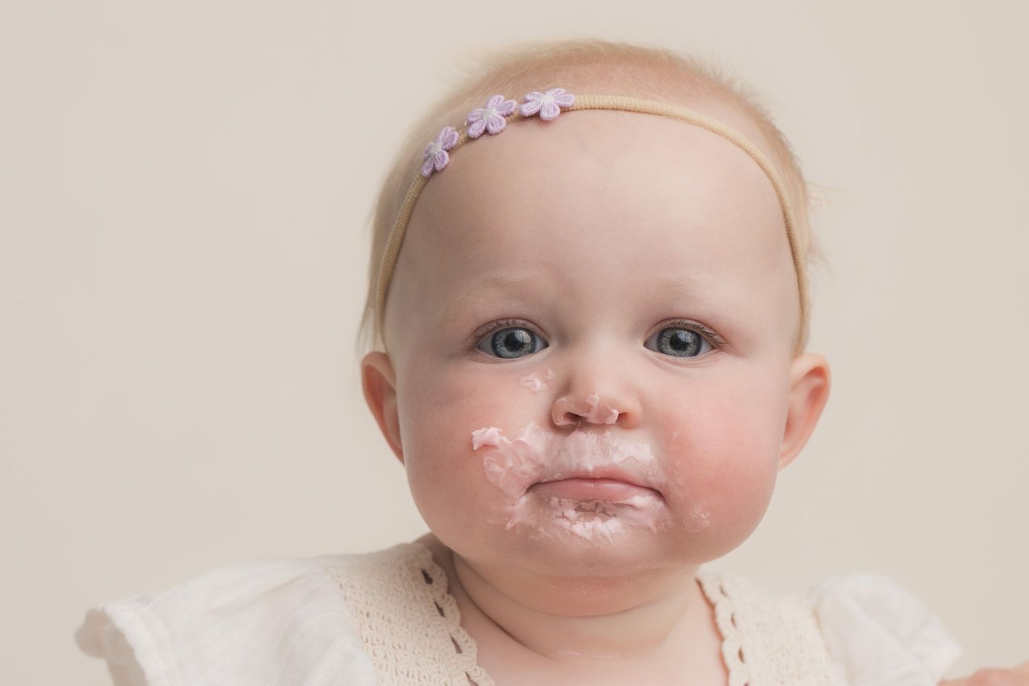 Cute blonde baby girl with food on her face wearing a pink floral headband and white outfit.