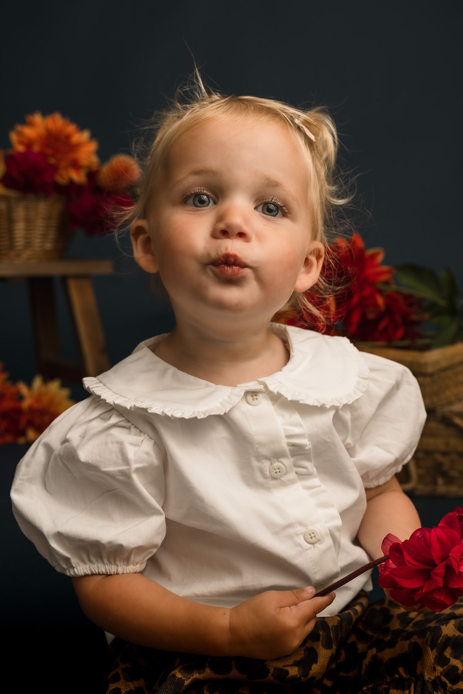 Adorable blonde toddler girl in white blouse making a kissy face, surrounded by colorful autumn flowers.