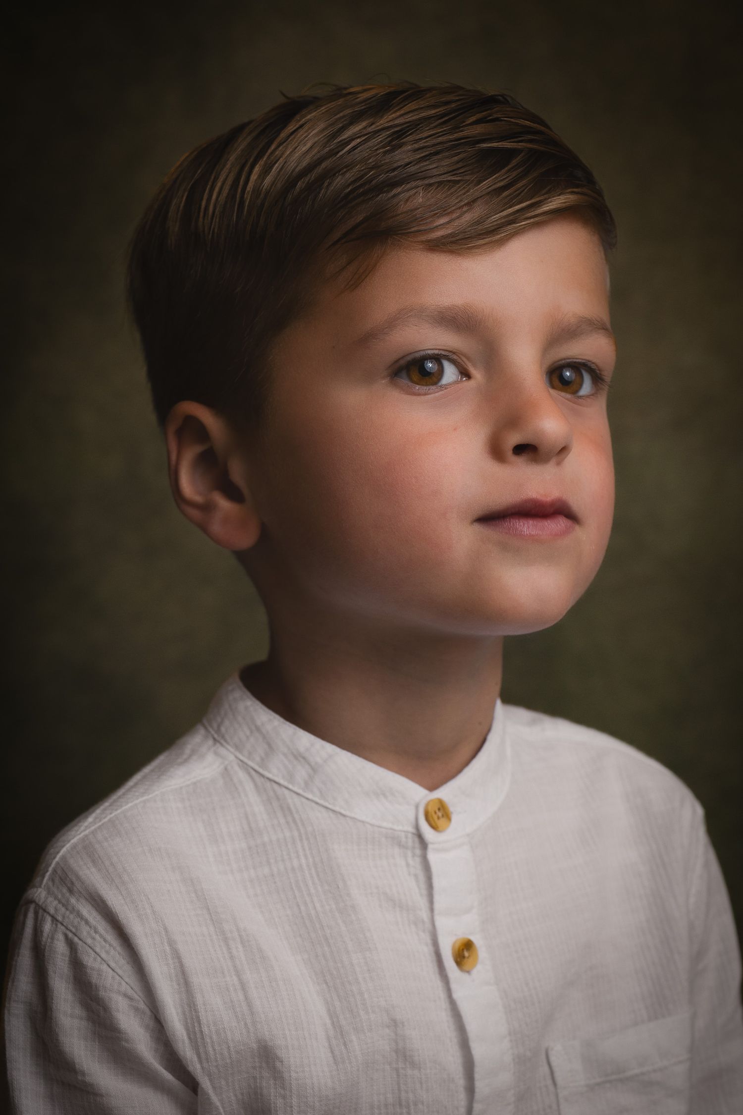 Young boy in white linen shirt with gold buttons poses for a classic studio portrait against dark background.