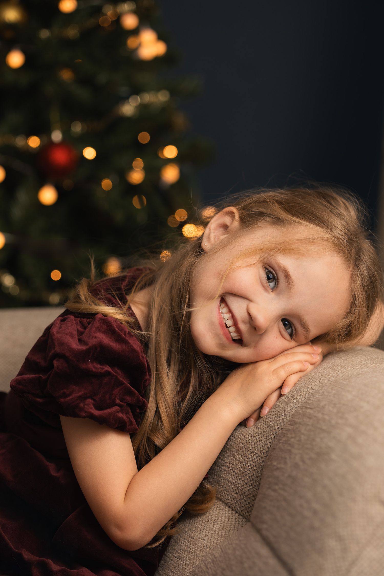 Lachend meisje tijdens een kerstfotosA smiling young girl in a burgundy velvet dress rests on a sofa in front of a decorated Christmas tree.