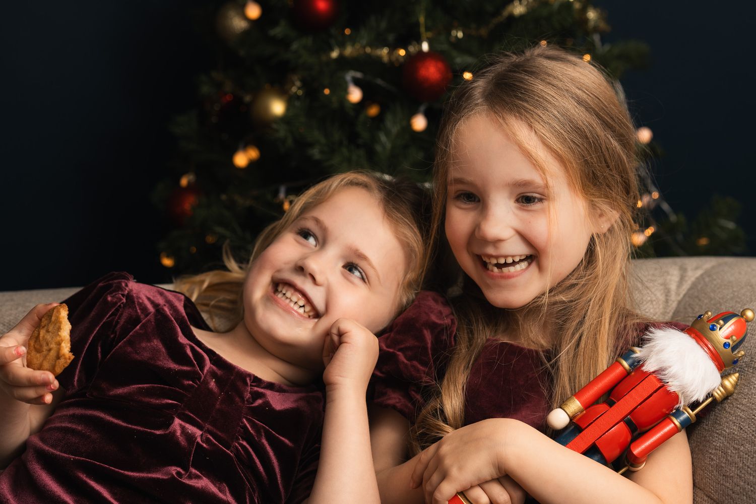 Two smiling girls in velvet dresses pose by a Christmas tree, one holding a nutcracker toy.