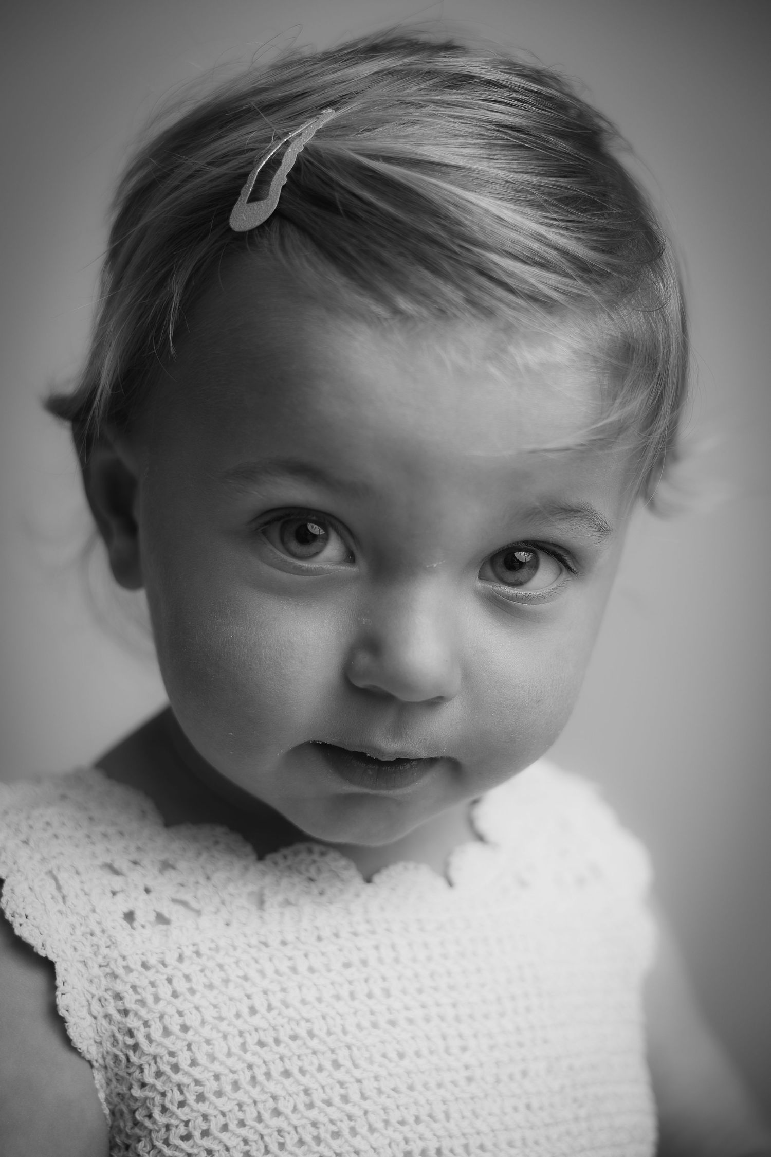 Zwart-wit kinderportret van een jonge peuter in een witte gebreide jurk met een subtiel haarspeldje.
