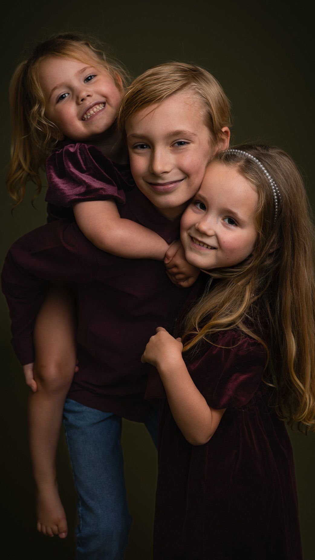 Three smiling young sisters in burgundy outfits pose together for a professional studio portrait photo.