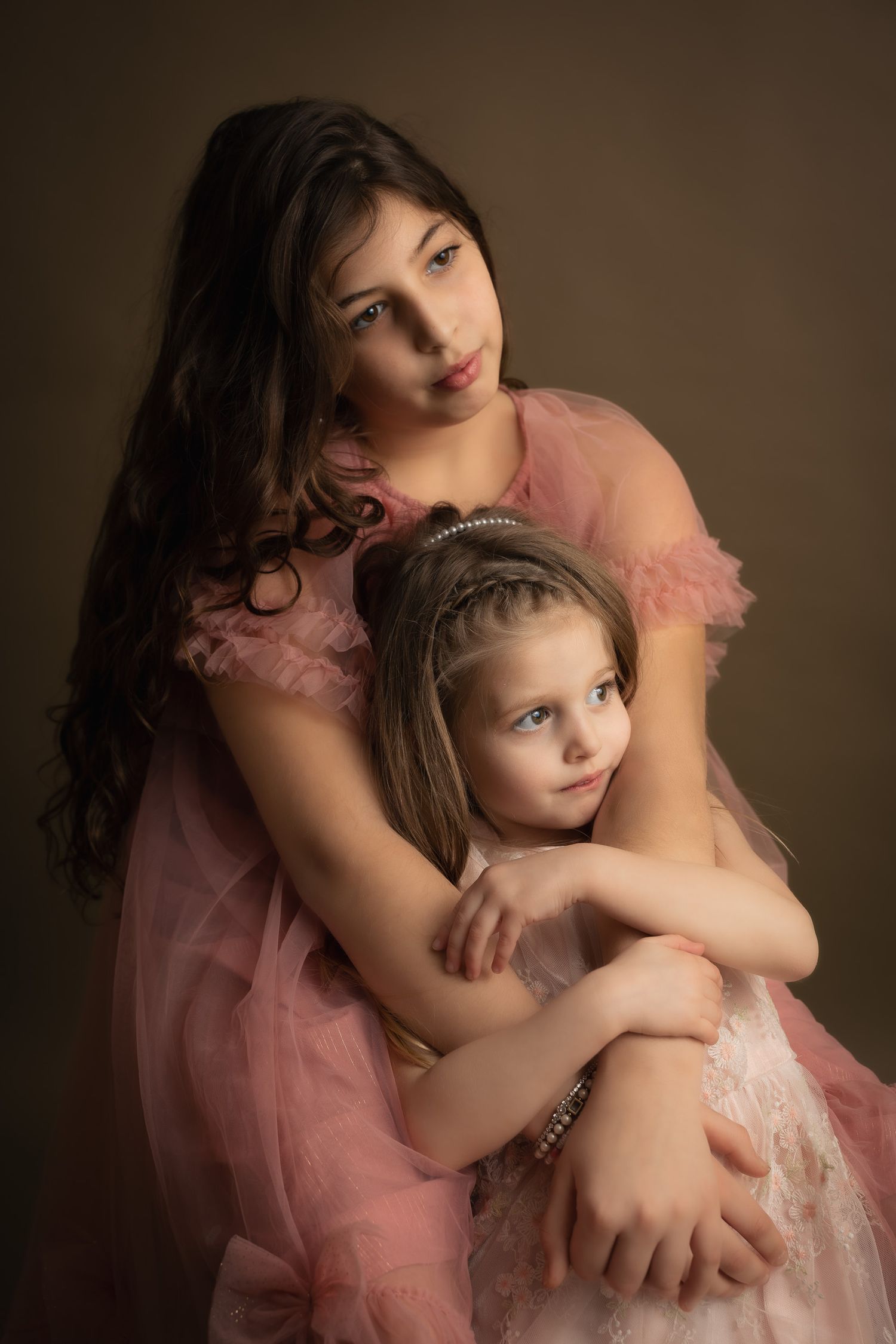 Two girls in pink tulle dresses pose together in a studio portrait, the older girl embracing the younger one.