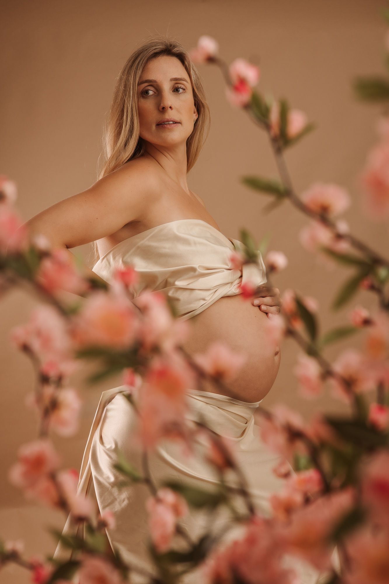 Zwangere vrouw in ivoorkleurige satijnen doek tussen zachtroze bloemen tijdens zwangerschapsfotografie