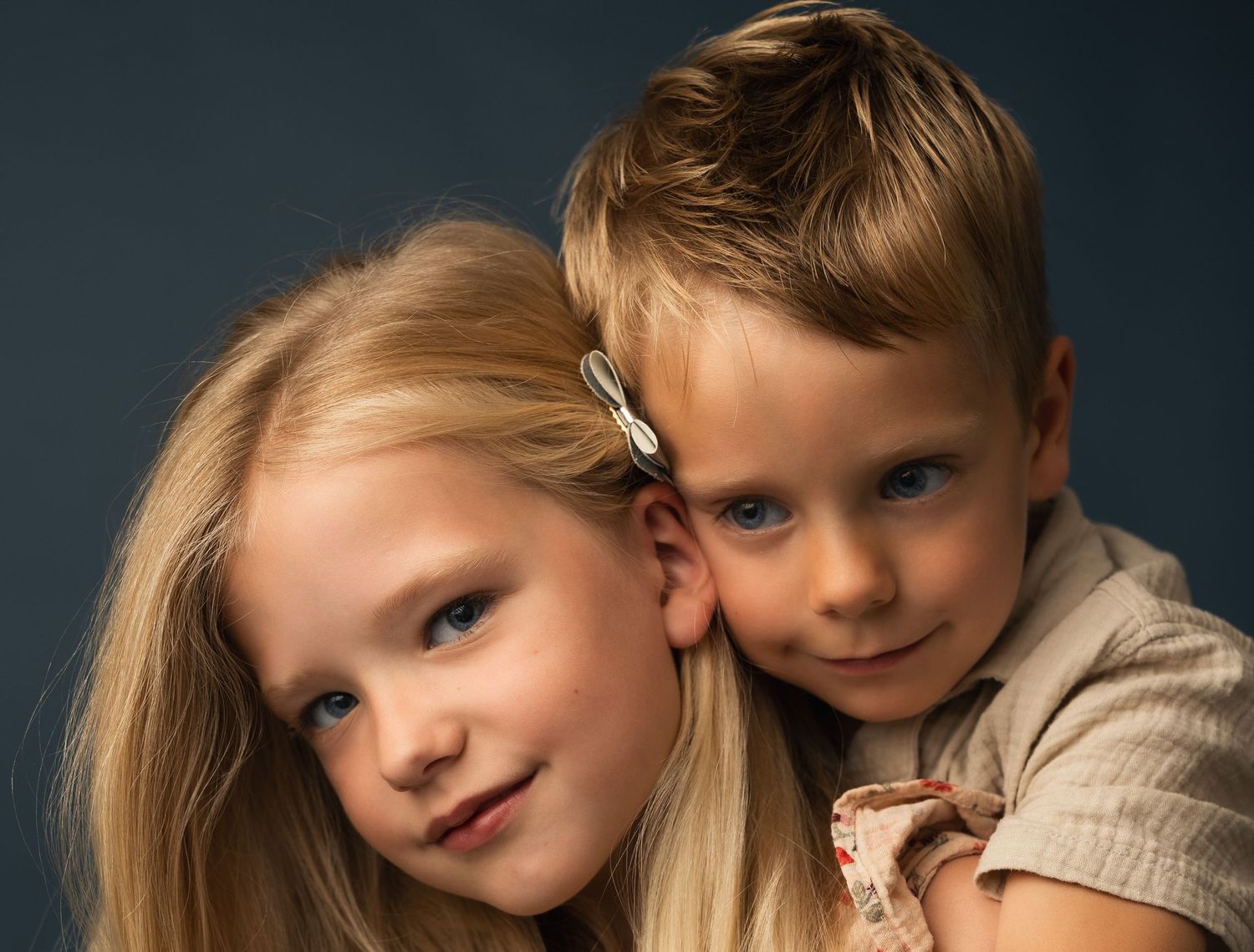 Two blonde children posing together for a studio portrait, a girl and boy smiling against a dark blue background.