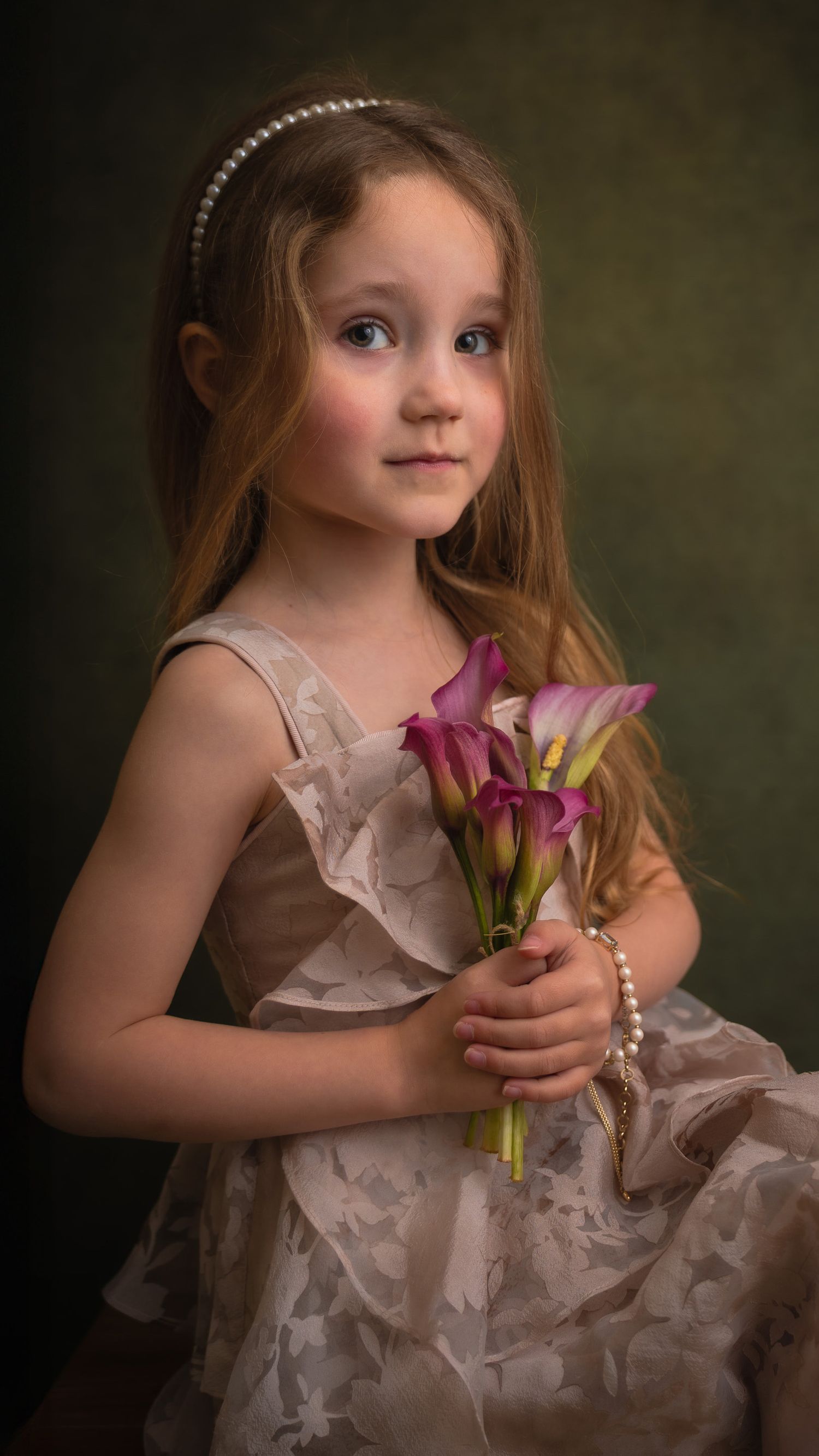 Young girl in elegant dress holding pink calla lilies, wearing pearl bracelet and headband, posed for formal portrait in photostudio Assen.