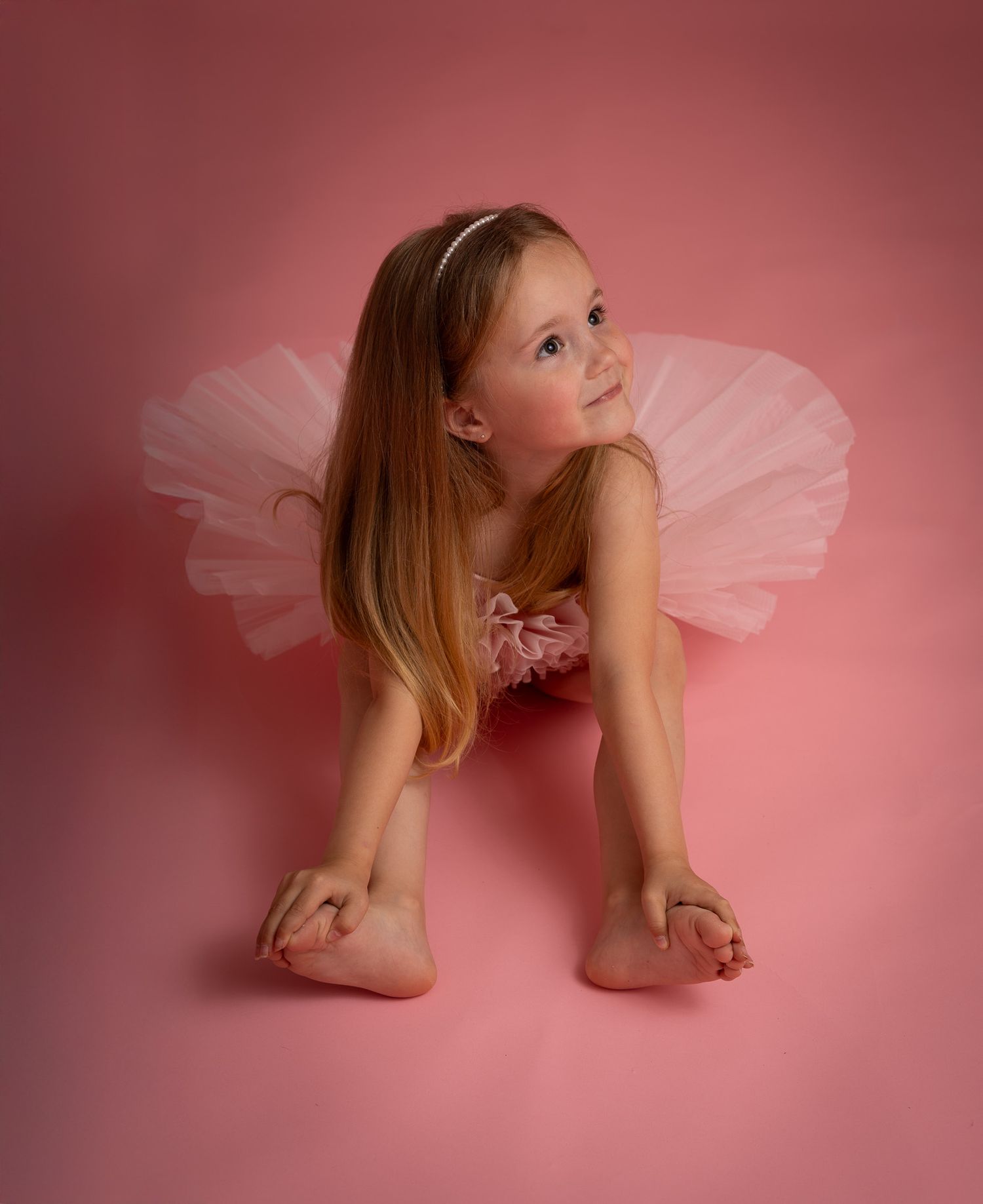 Young girl in pink tutu ballet costume poses gracefully on pink background, looking up with long hair flowing.