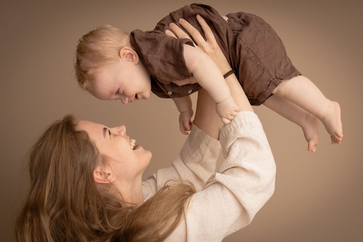 A joyful mother lifts her smiling toddler overhead against a warm beige studio background.