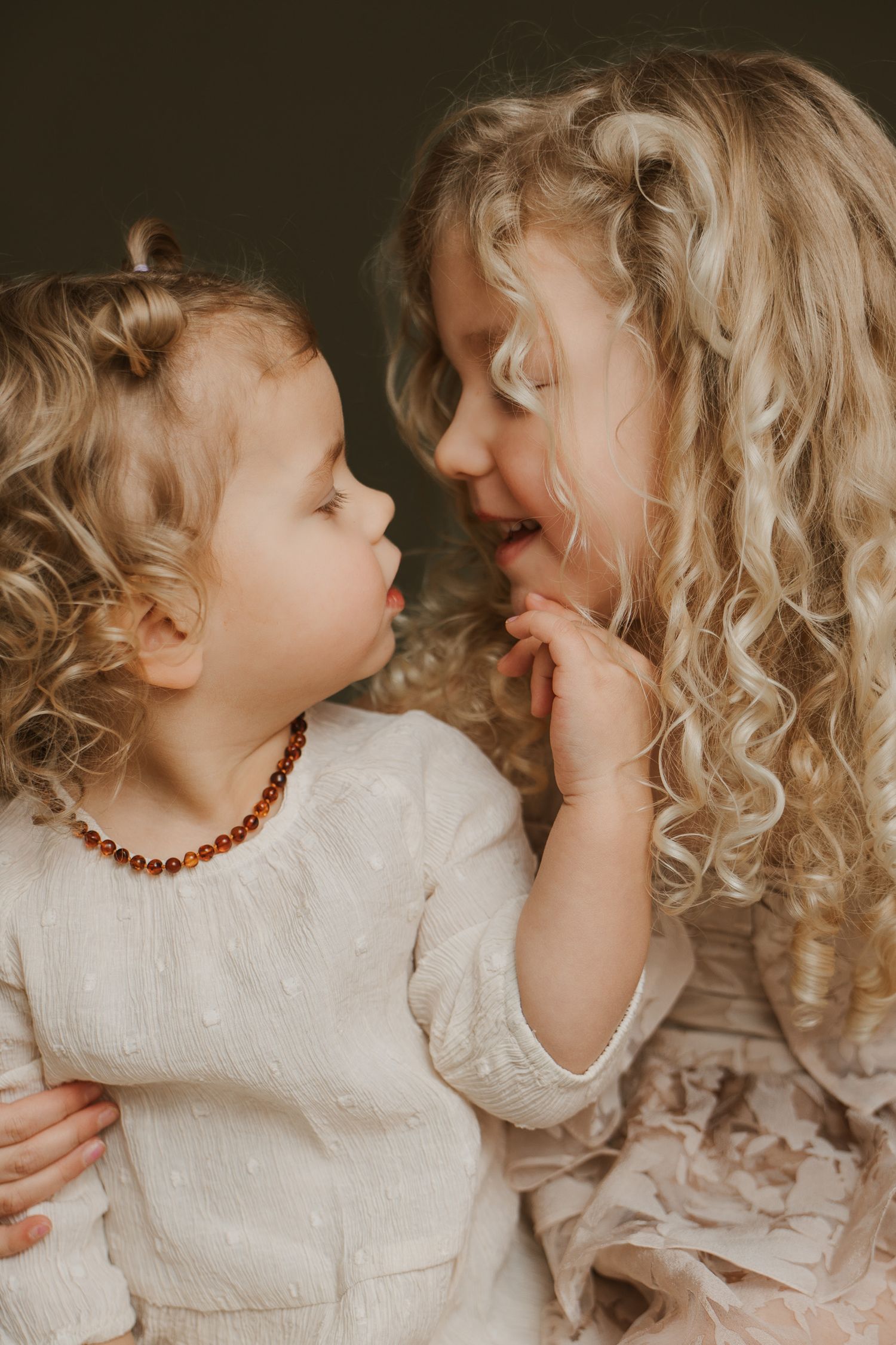Two young girls with curly blonde hair share a sweet moment, one wearing an amber bead necklace, dressed in soft cream outfits.