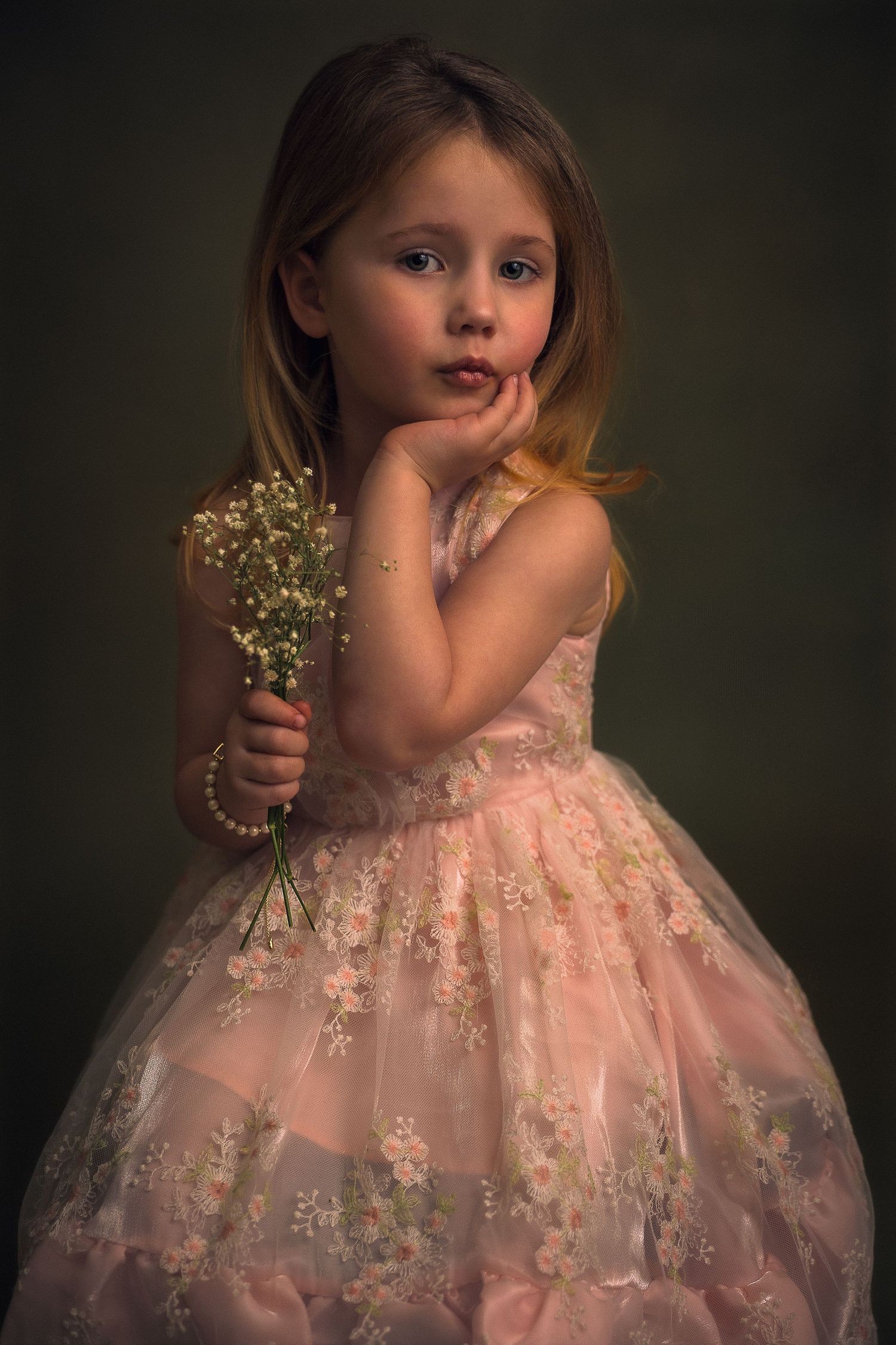 Young girl in pink floral dress holding white wildflowers, posing thoughtfully against dark background.