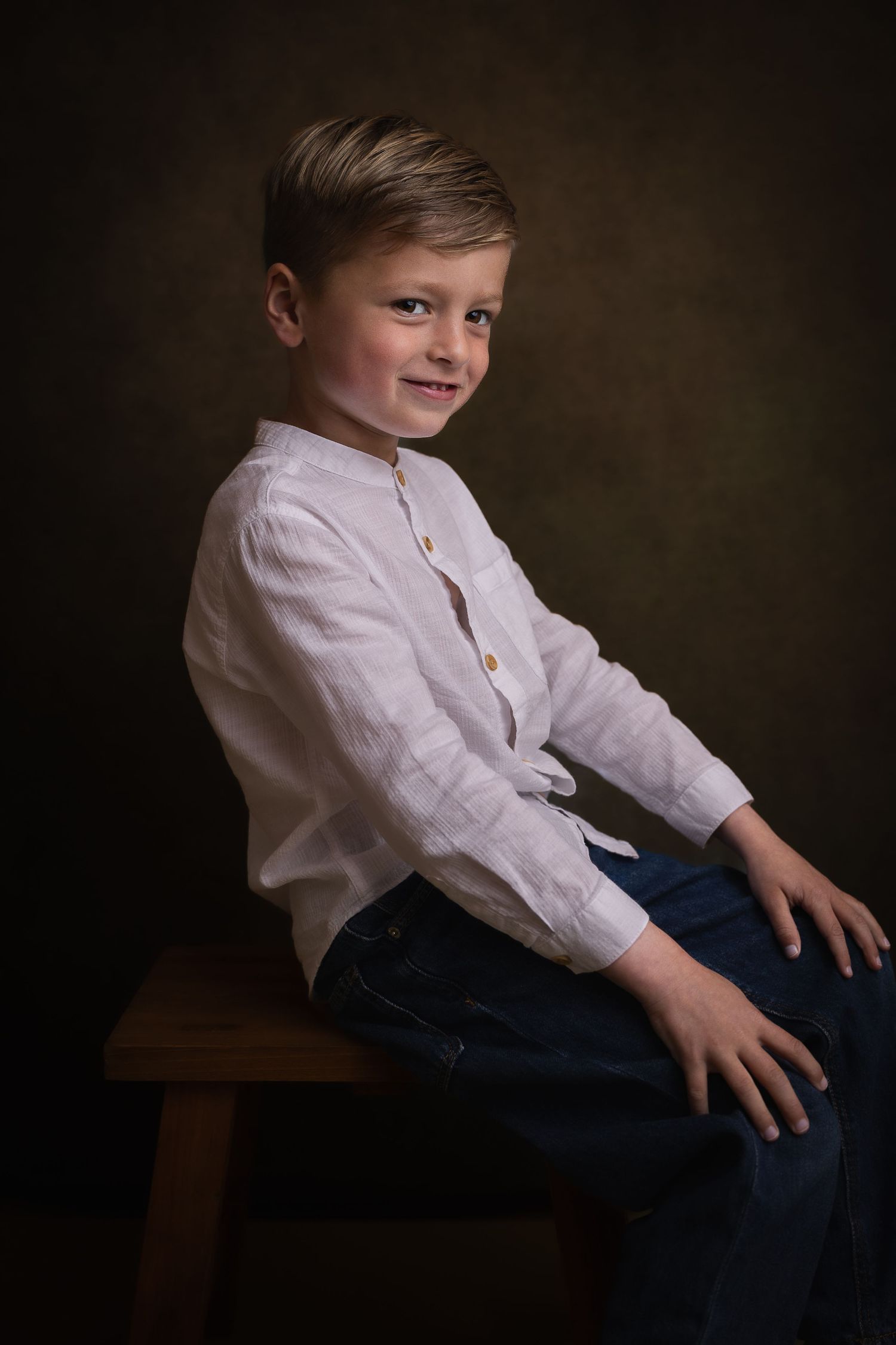 Klassiek kinderportret van een glimlachende jongen in een wit linnen overhemd op een houten kruk.
