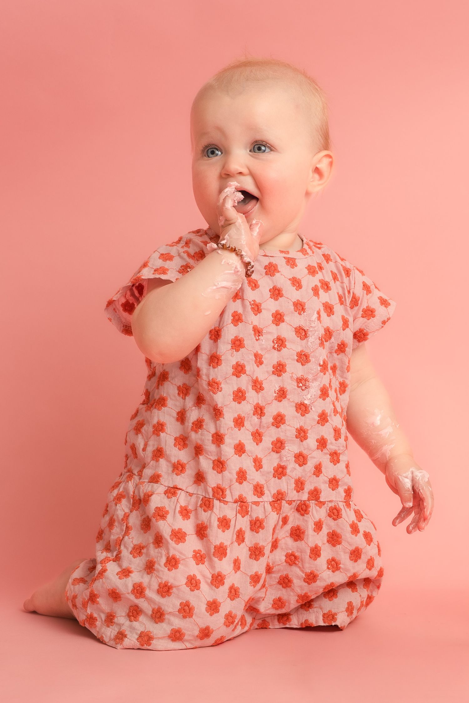 Adorable baby girl in a floral orange dress kneeling on a pink background, chewing on her fingers.