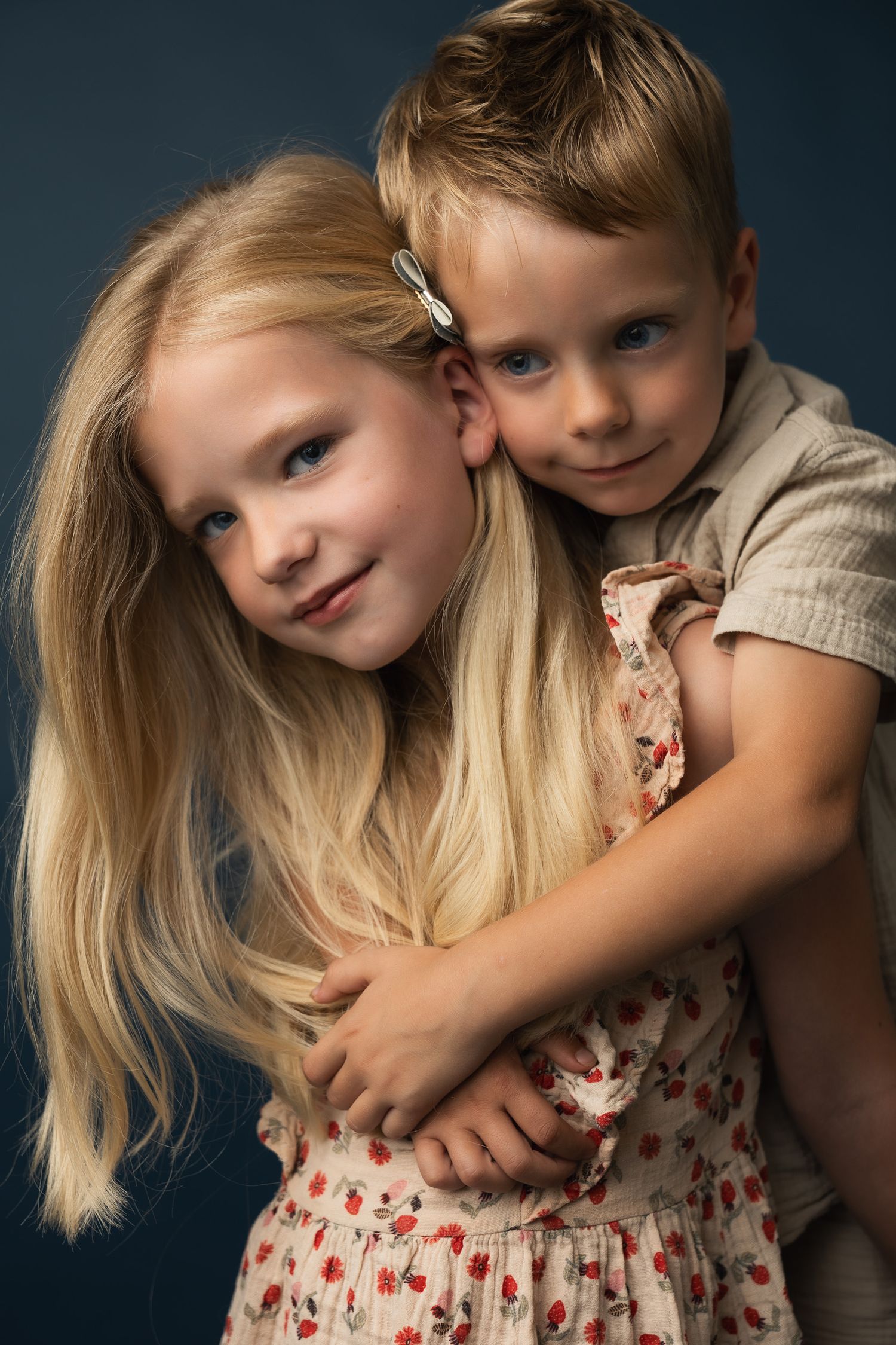 Two blonde children hugging warmly against a dark blue background in a studio portrait photo.