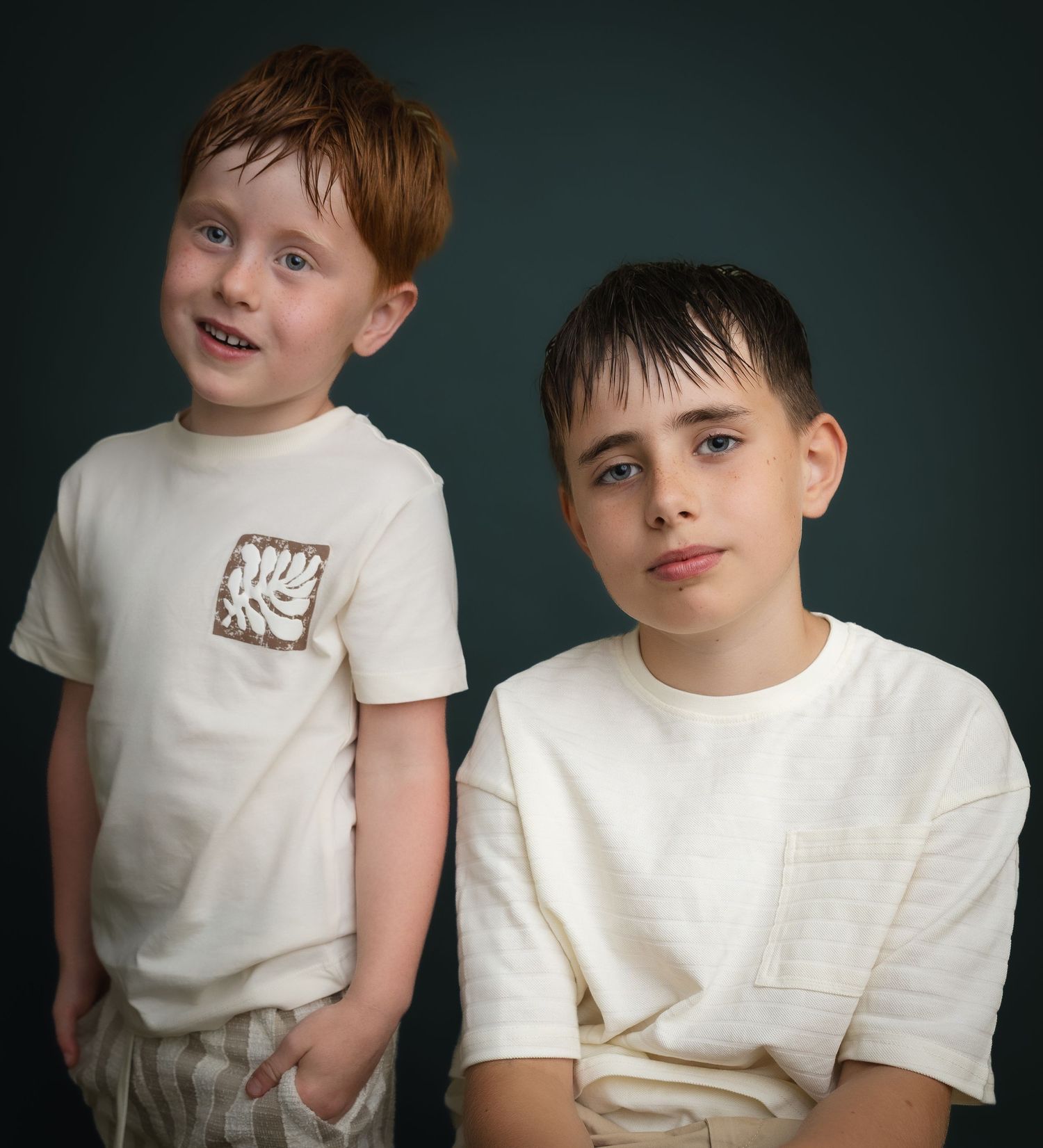 Two boys in white t-shirts posing for a portrait, one with red hair and one with dark hair, against a dark background.