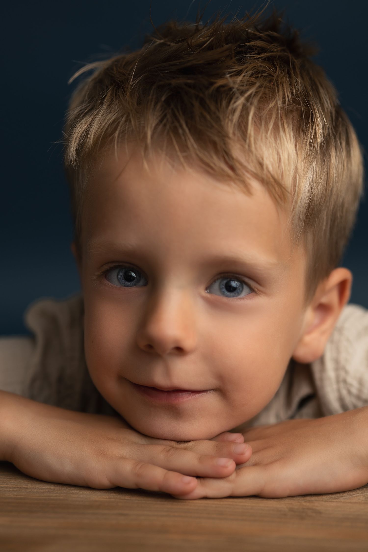 Adorable young boy with blue eyes and blonde hair resting his chin on folded hands on a wooden surface.