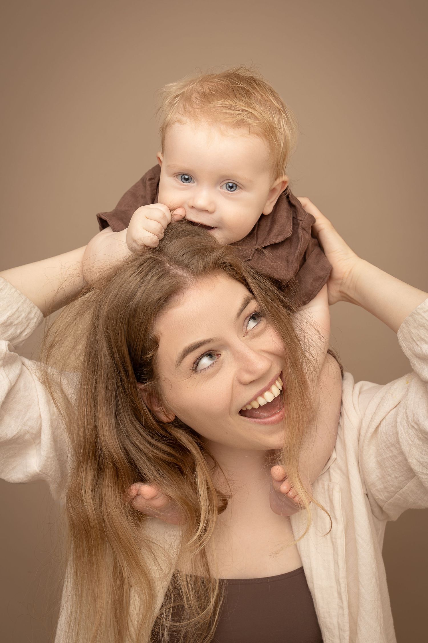 A joyful young mother holds her blue-eyed baby on her shoulders, both smiling against a beige background.