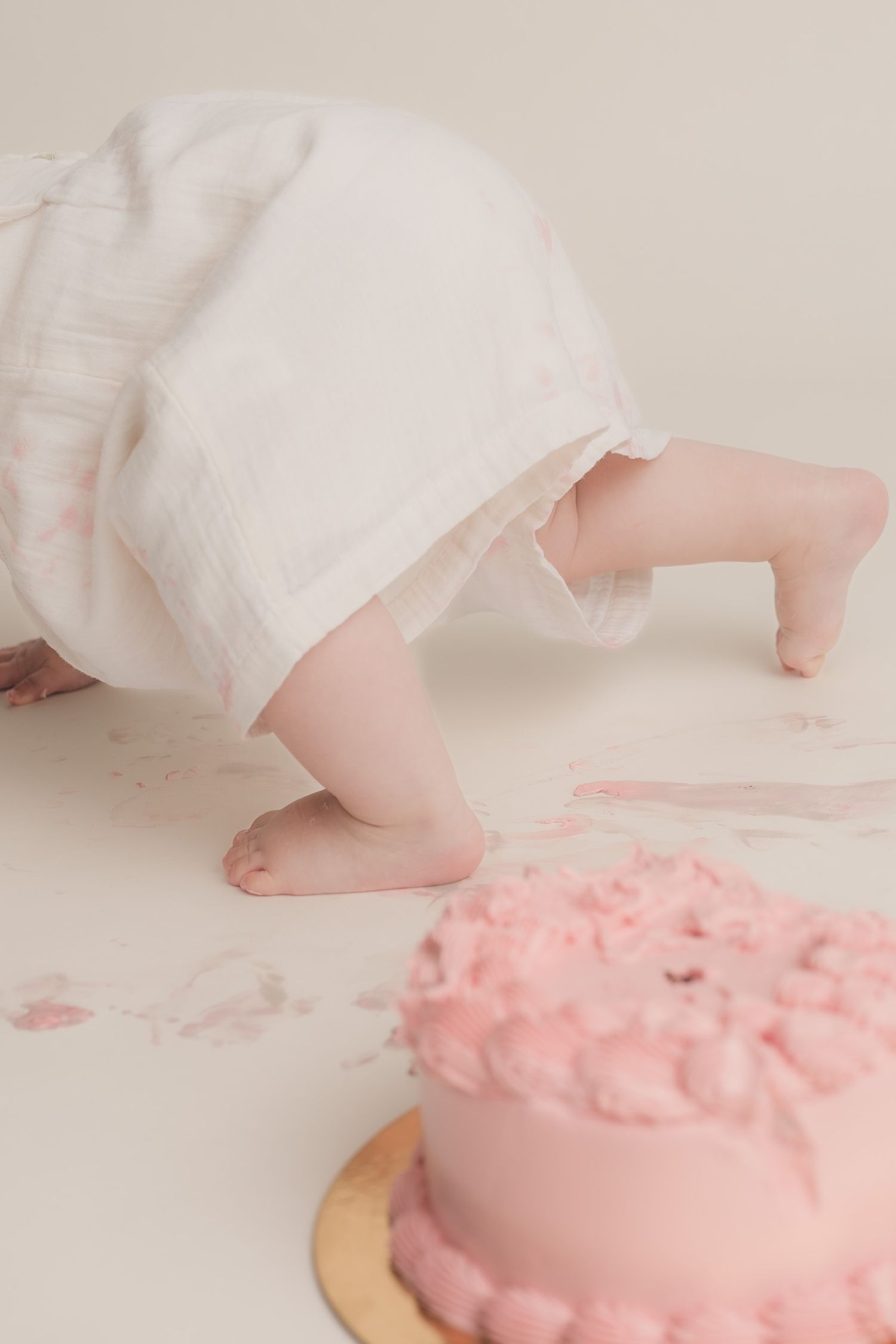 Baby in white dress crawling near a pink smash cake on white background during first birthday photoshoot.