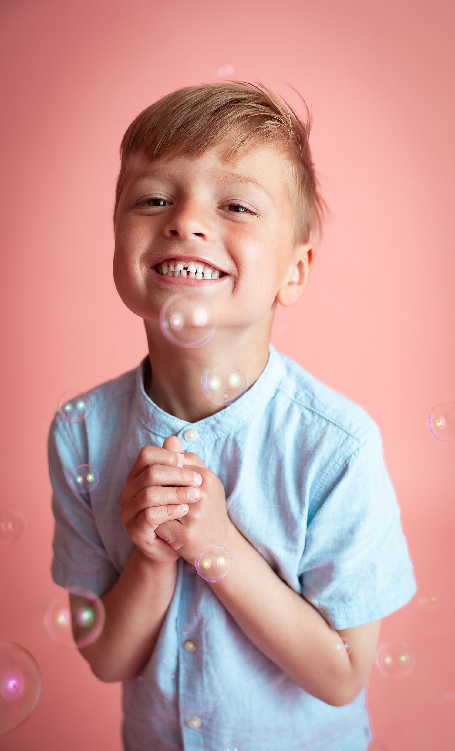 A happy young boy in a light blue shirt smiles while playing with soap bubbles against a pink background.