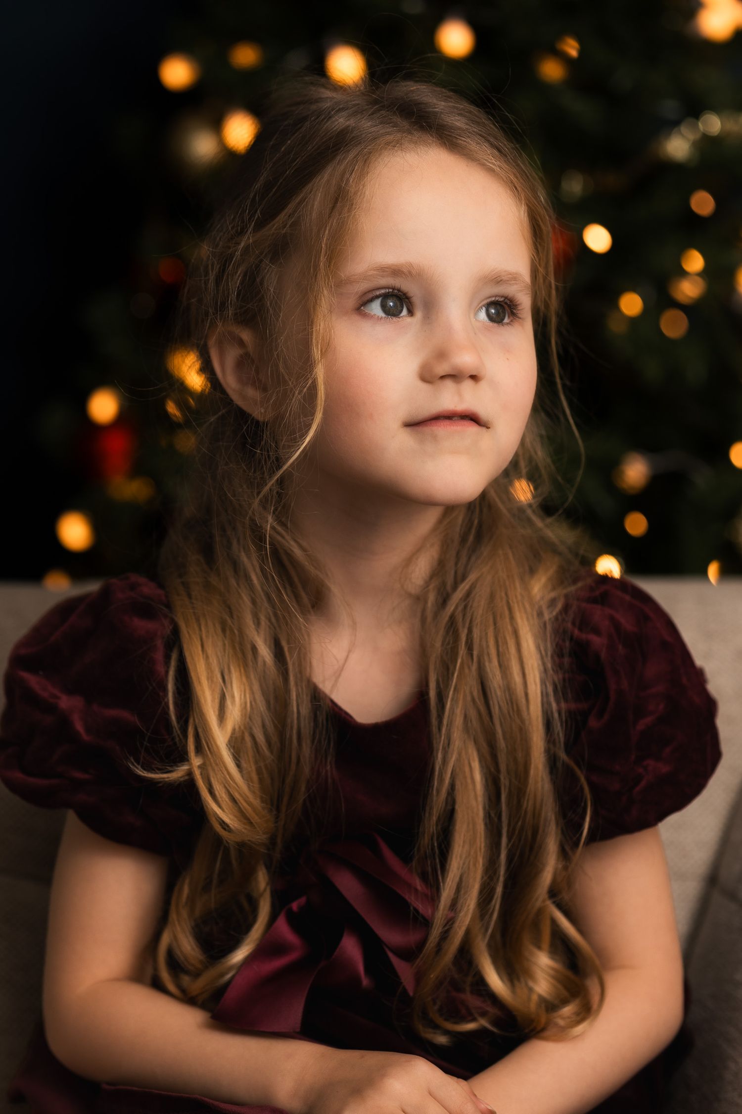 Rustig kinderportret inYoung girl in burgundy velvet dress with long hair poses before a glowing Christmas tree in festive holiday portrait.