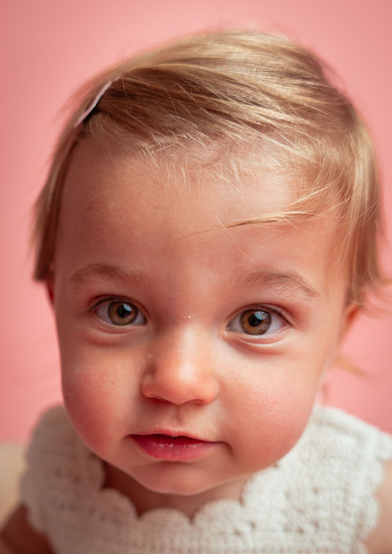 Close-up portrait of a blonde toddler girl with brown eyes wearing a white dress against a pink background.
