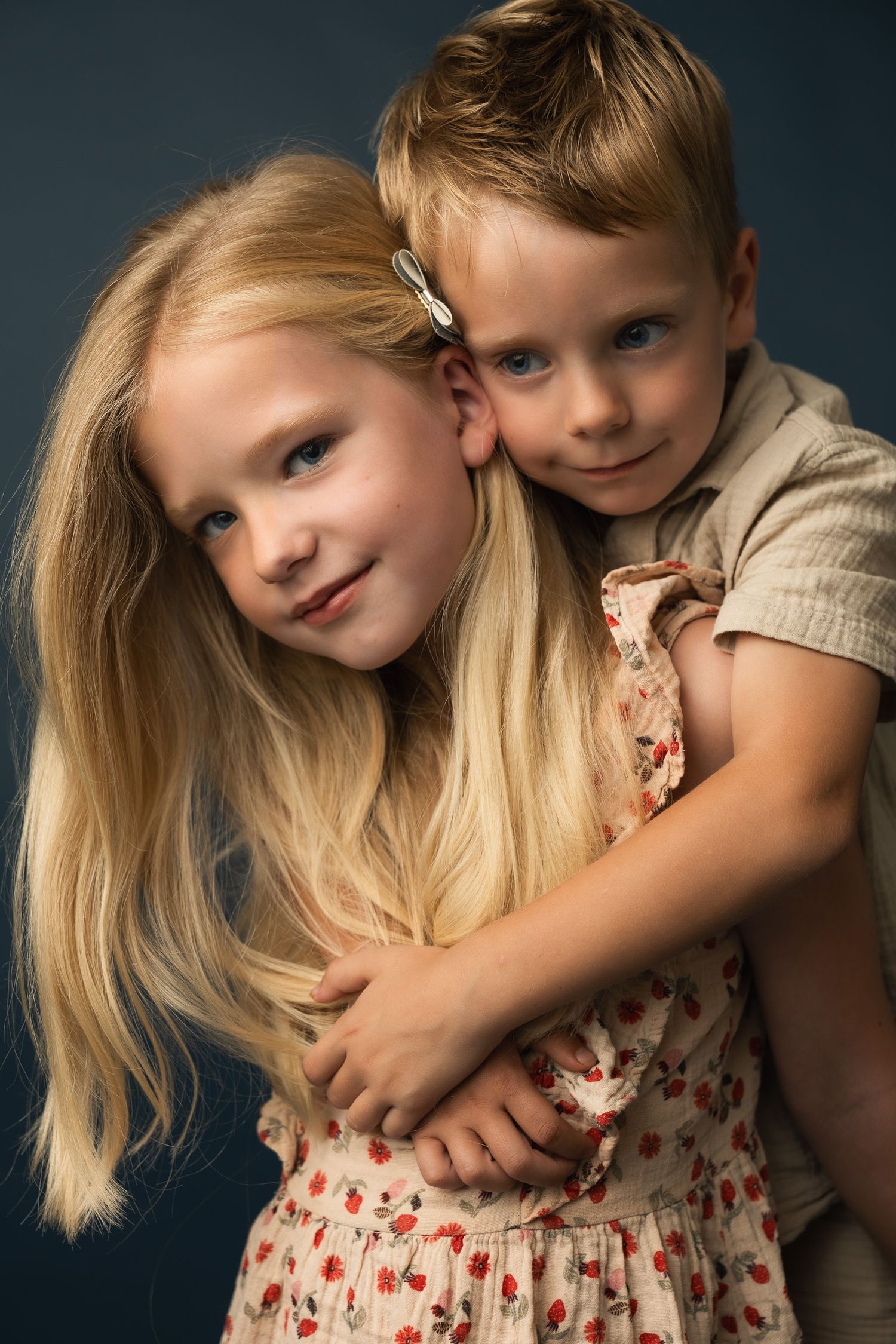 Two blonde children hugging in a studio portrait, girl in floral dress with long hair, boy smiling behind her.