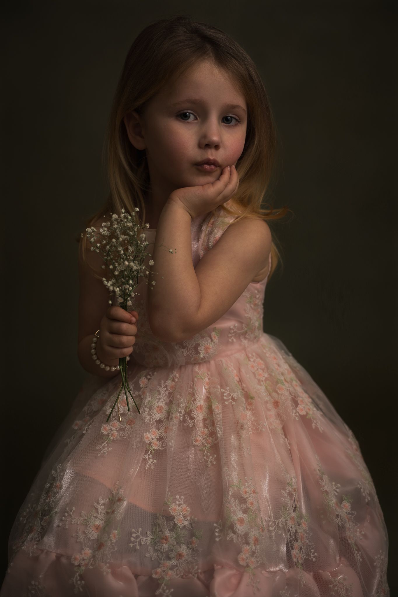 portret van een meisje in een roze jurk met bloemen tegen een donkergroen achtergrond met een bosje gipskruid in haar hand