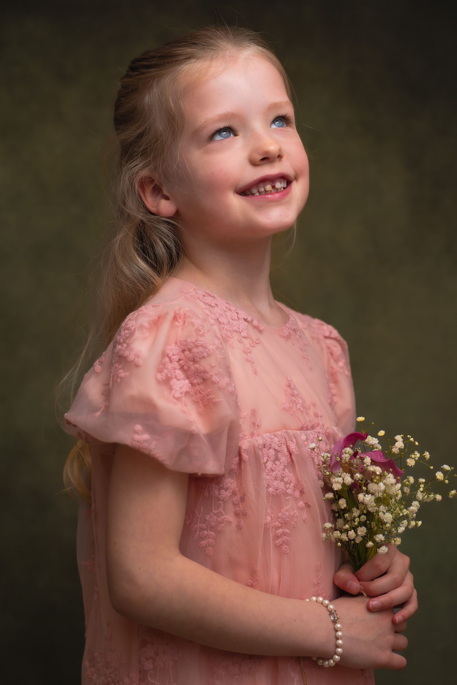 Young blonde girl in pink lace dress holds wildflowers, smiling upward, wearing pearl bracelet, studio portrait made in photostudio Assen.