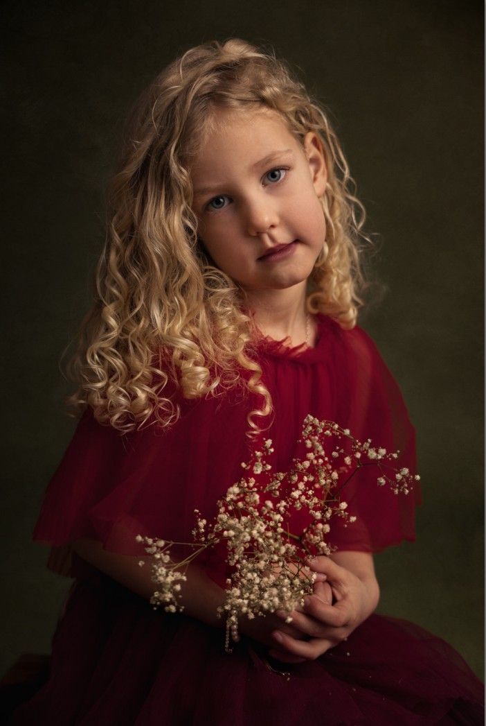 Young girl with curly blonde hair in red dress holding delicate baby's breath flowers, elegant portrait photography.