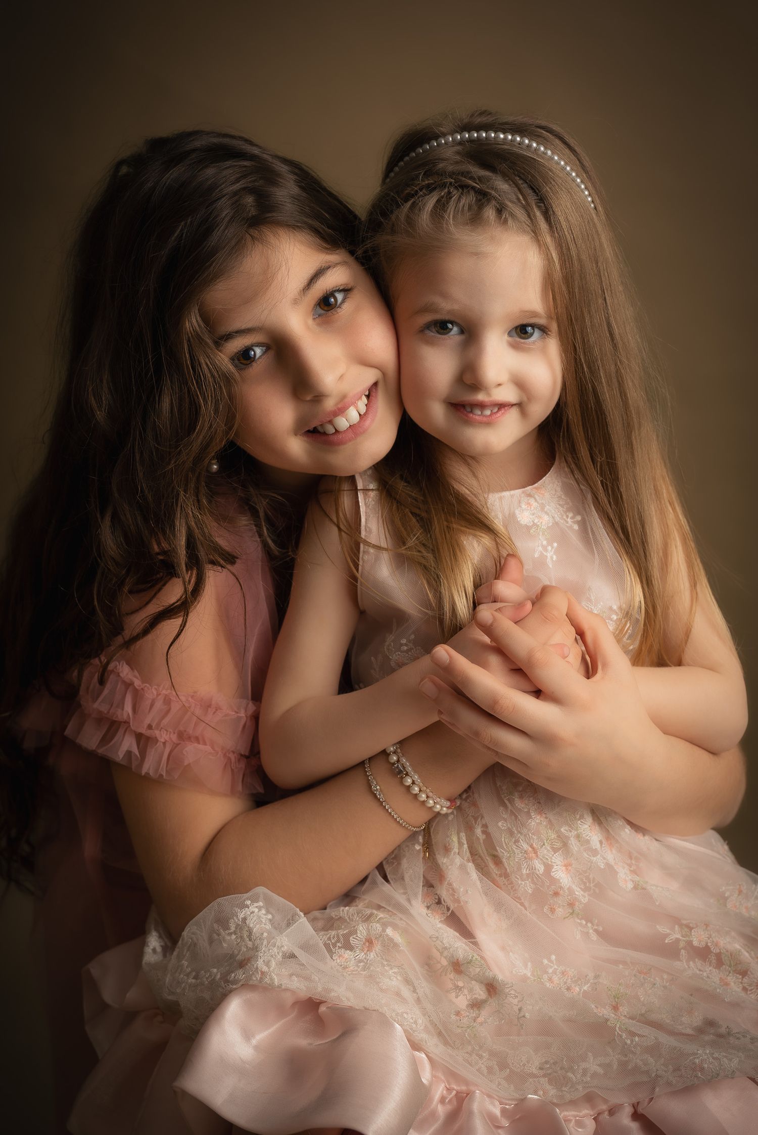 Two young sisters in pink dresses share a warm embrace, smiling for a professional portrait photo.