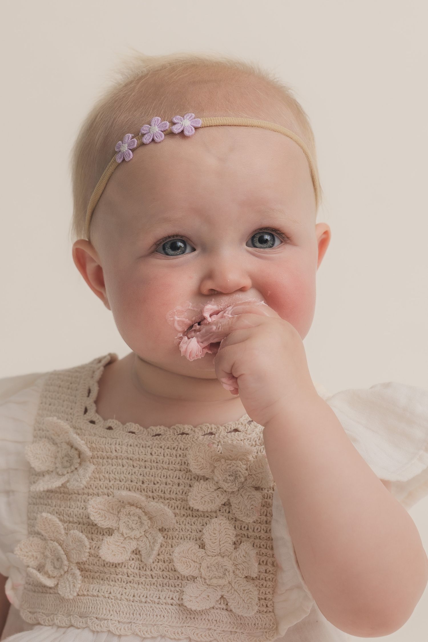 Blue-eyed baby girl wearing a floral headband and lace dress, eating with her hand during a portrait session.