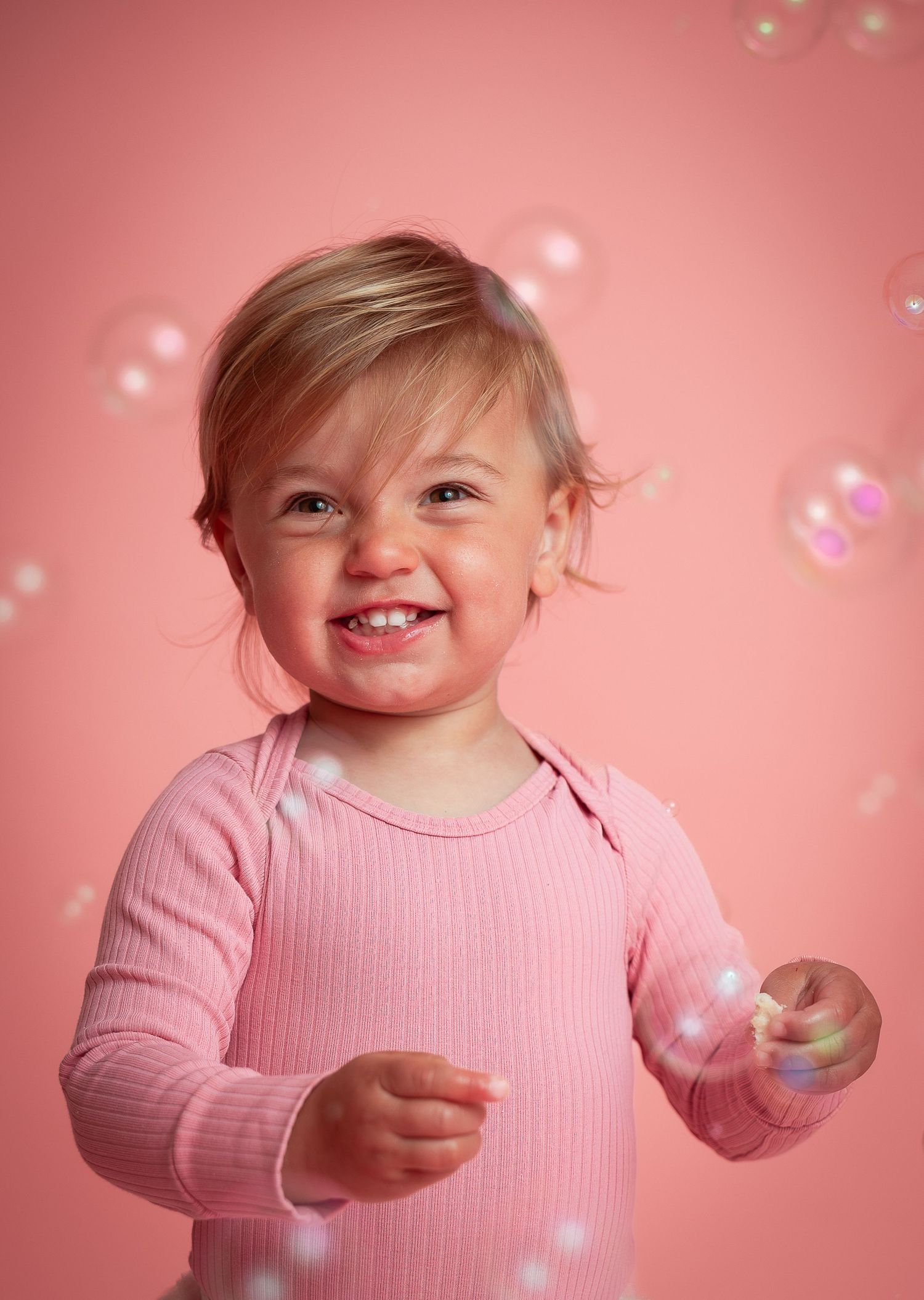 A smiling toddler in a pink ribbed top plays with soap bubbles against a soft pink background.