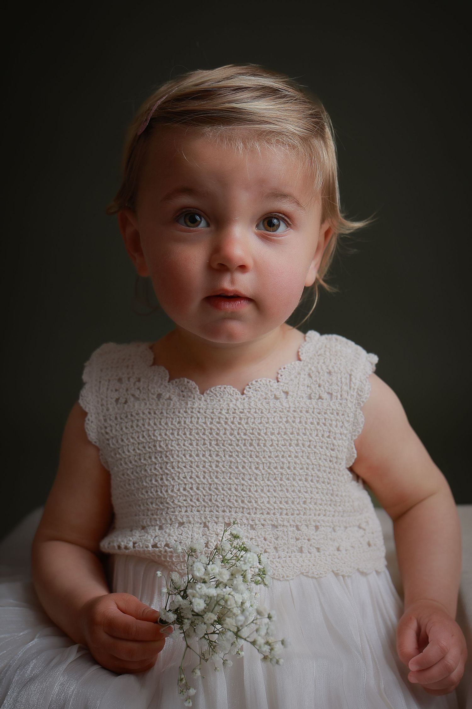 Sweet baby girl in white crochet dress holding delicate white flowers in a professional studio portrait.