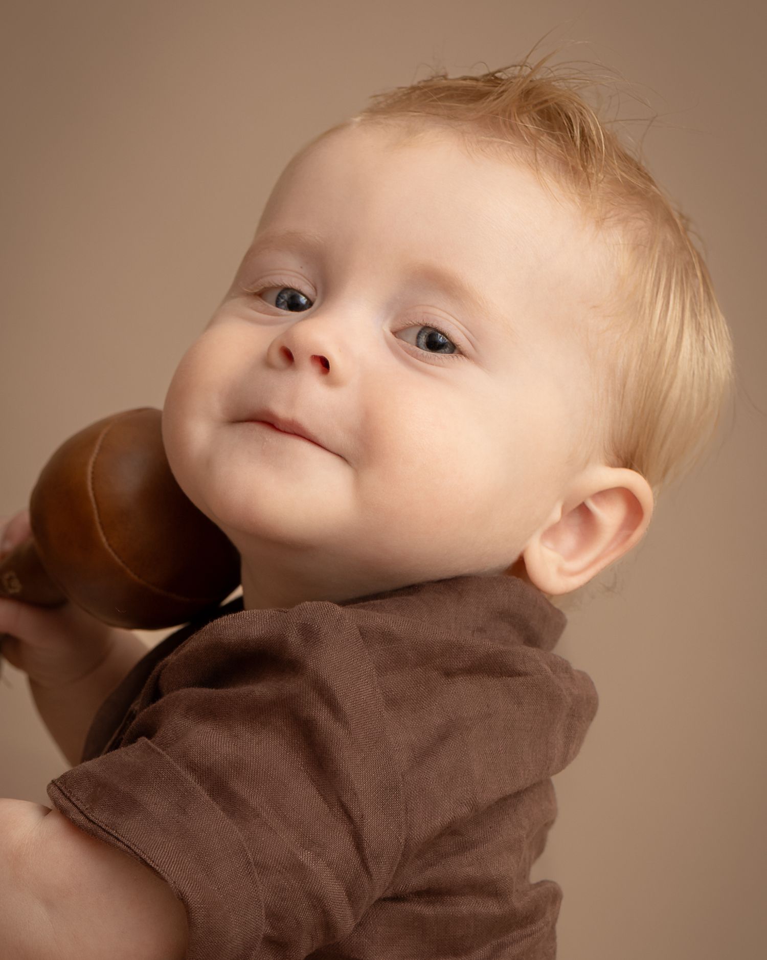 Adorable blue-eyed baby with blonde hair holding a brown wooden toy, wearing a brown outfit against a beige background.