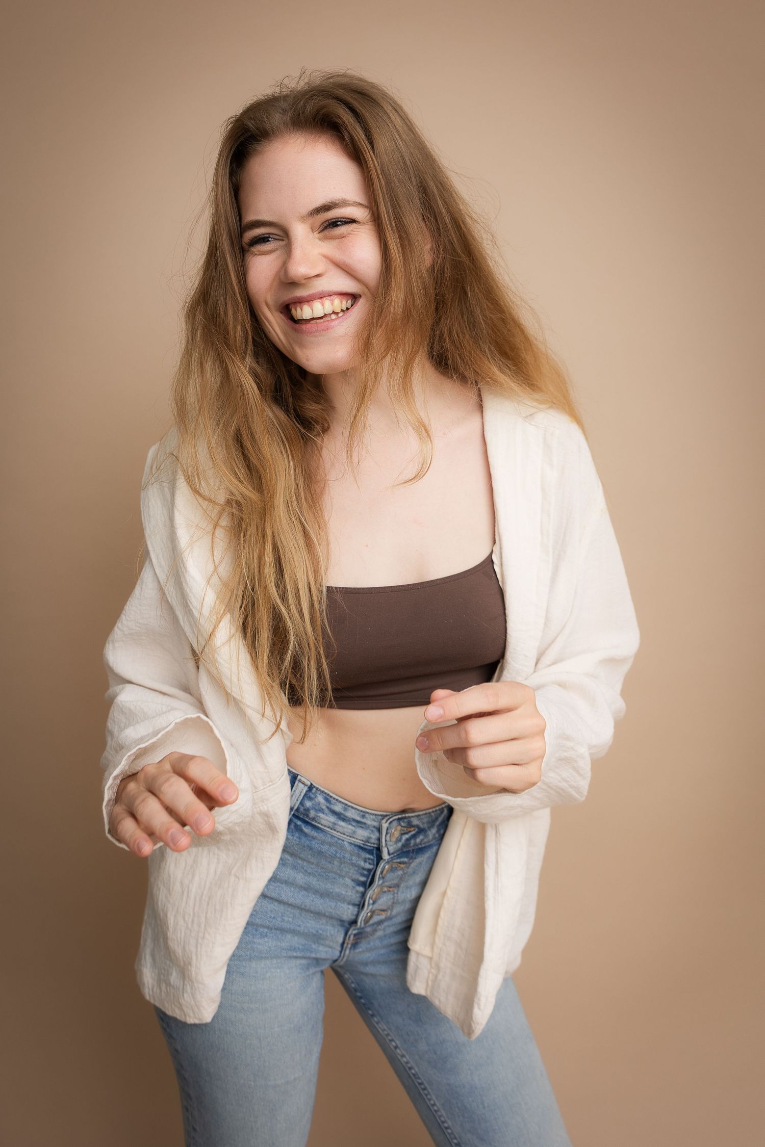 Young woman laughing joyfully, wearing white shirt and brown crop top with jeans, posing against beige background.