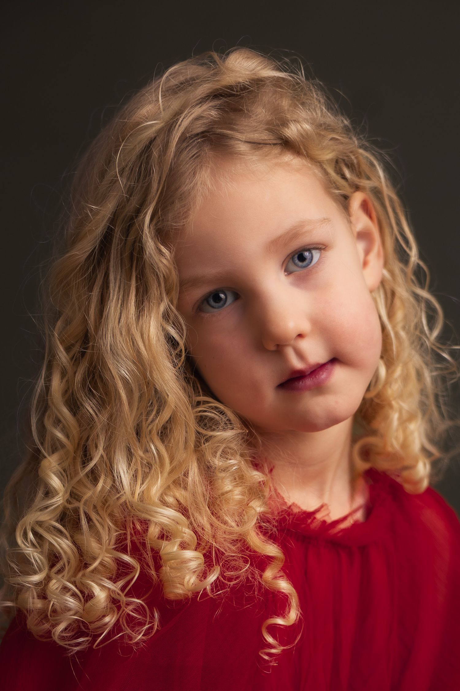 Young girl with long curly blonde hair and blue eyes wearing a red dress, posed against a dark background.