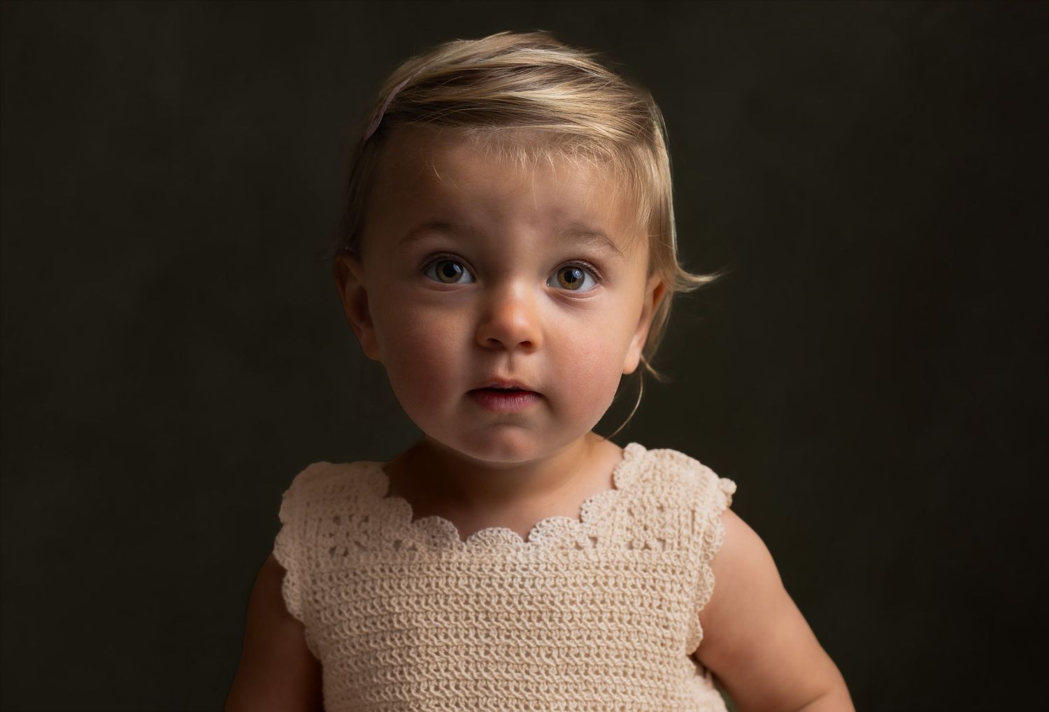 Adorable toddler girl with blonde hair wearing a cream crochet dress, posing for a professional portrait photo.