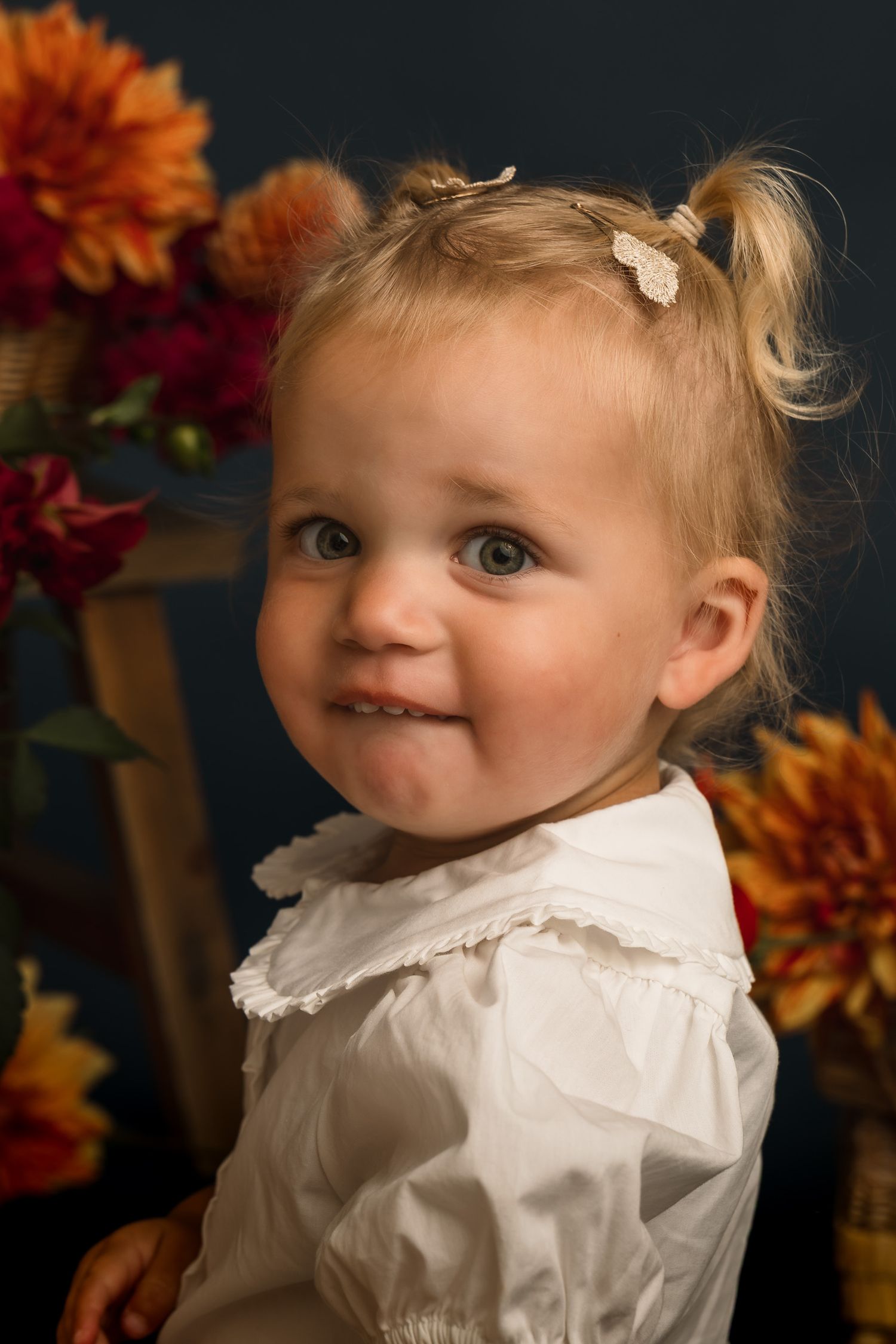 Adorable blonde toddler girl with pigtails smiling, wearing white dress, surrounded by autumn chrysanthemums.