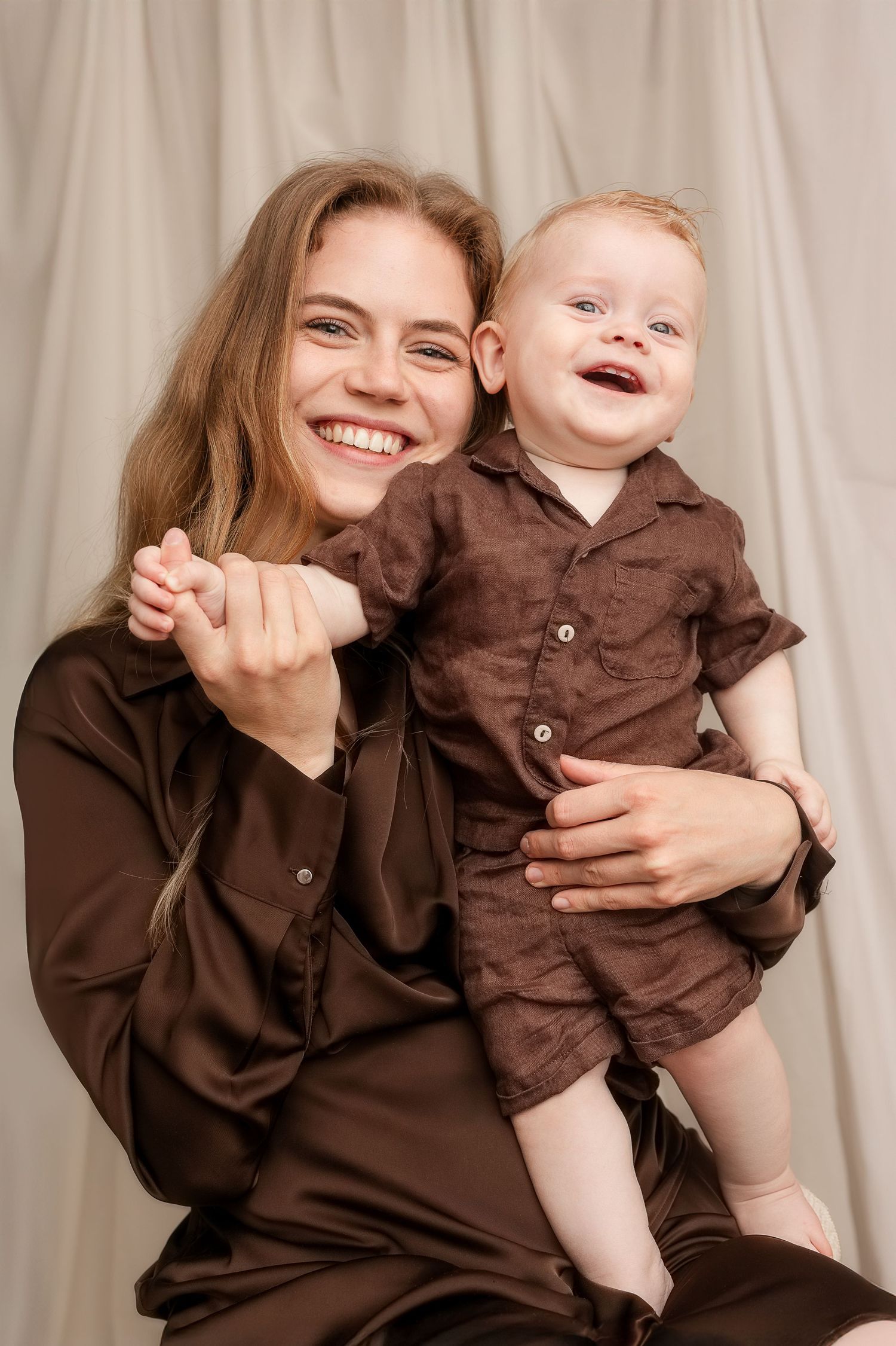 Smiling young woman in brown outfit holding a laughing baby dressed in matching brown clothes against neutral backdrop.
