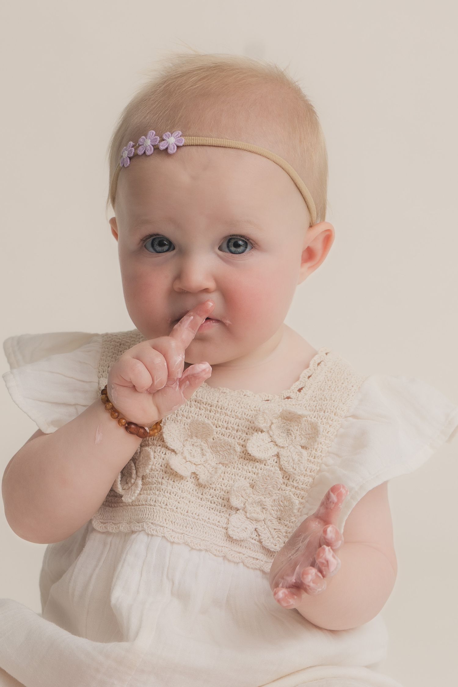 Adorable baby girl with blue eyes wearing a white crochet dress and purple floral headband, finger touching her lips.