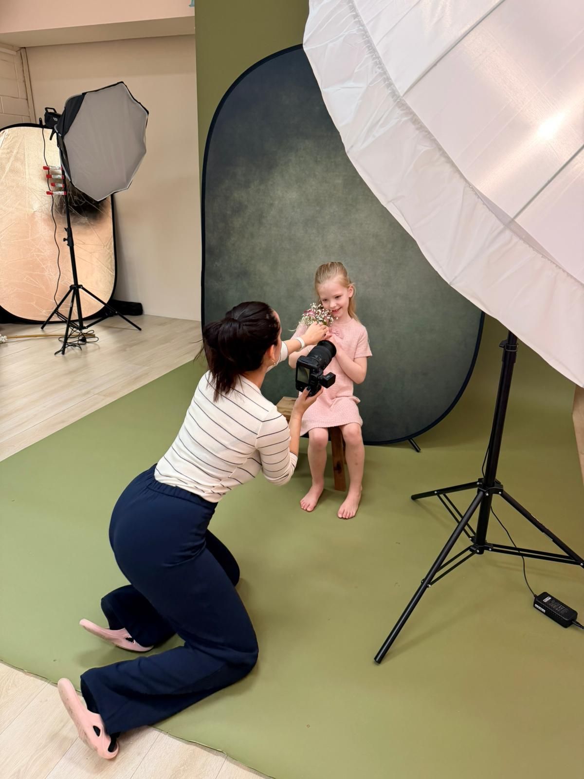 Fotograaf tijdens een kinderfotoshoot met een jong meisje in een roze jurk voor een donkere achtergrond.