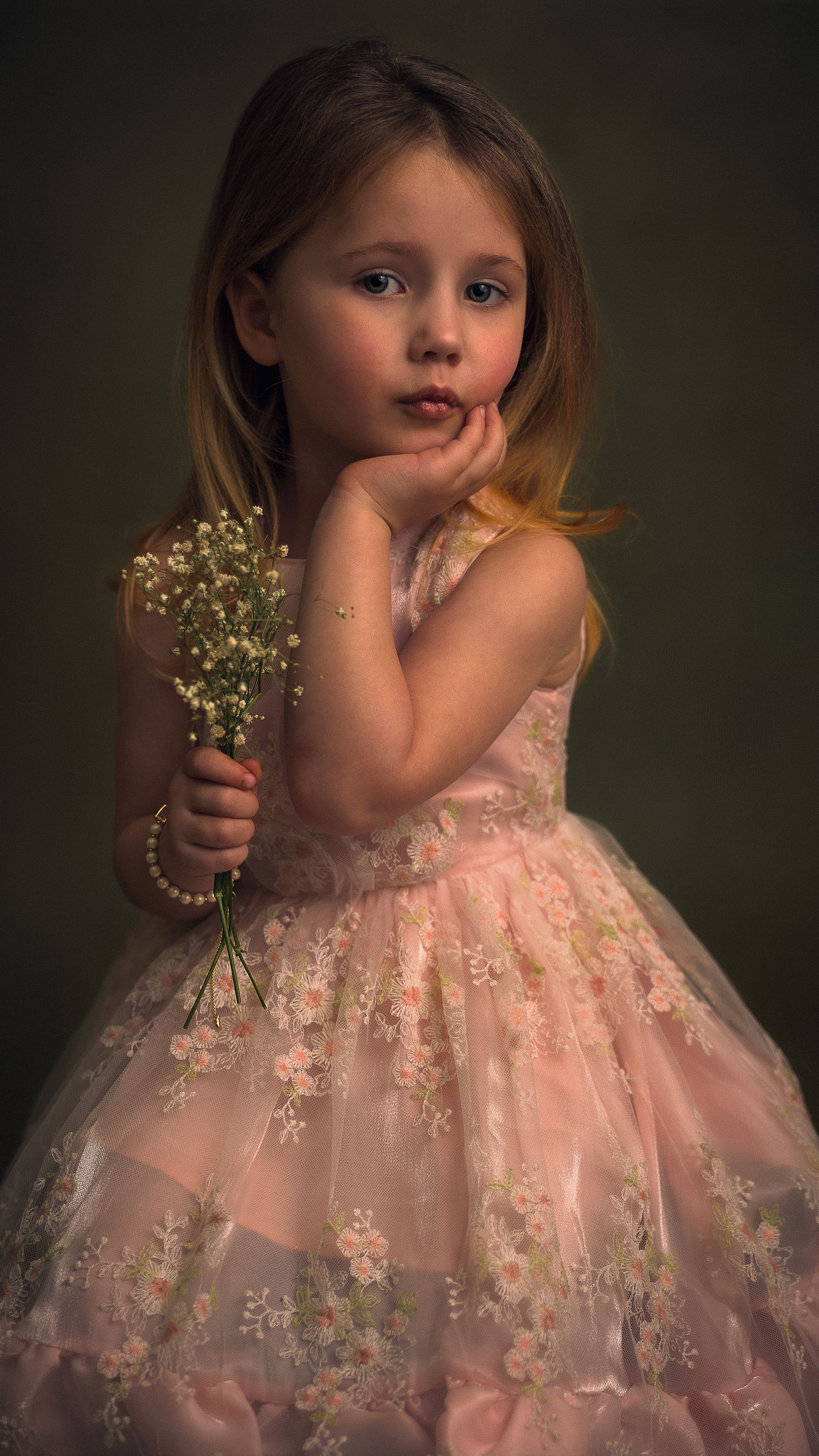 A young girl in a pink floral tulle dress holds white baby's breath flowers while posing thoughtfully.