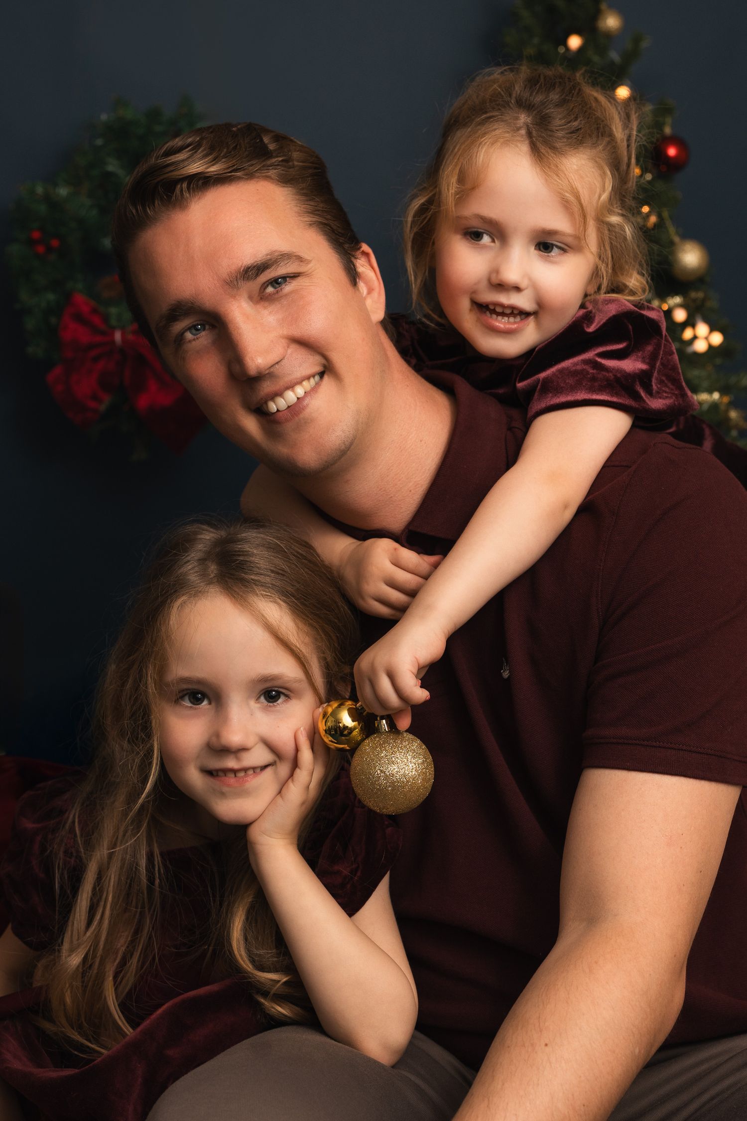 Father poses with two young daughters in matching burgundy outfits for a festive Christmas portrait.