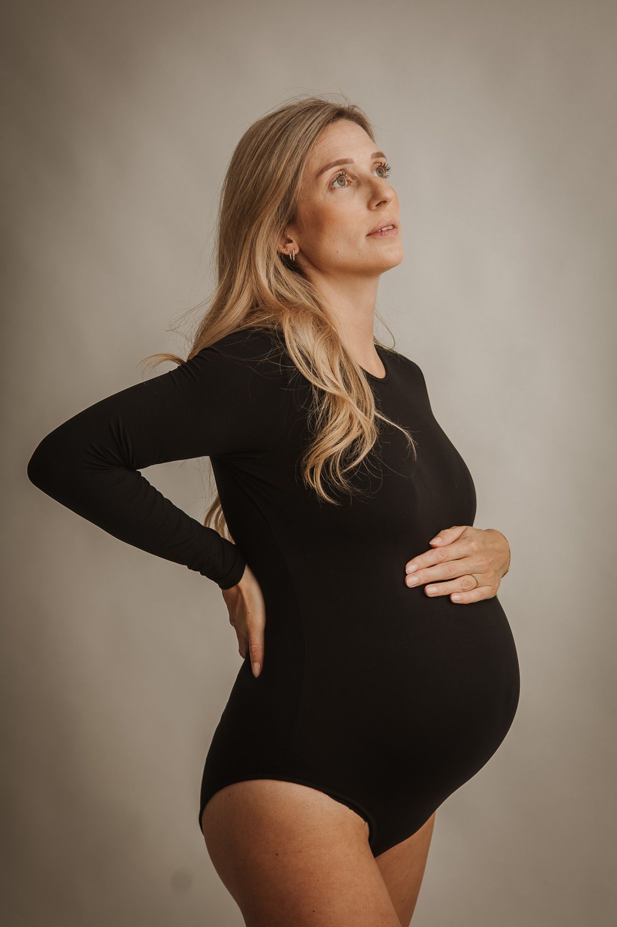Pregnant woman in black bodysuit cradling baby bump, posing against neutral background in maternity photo shoot.