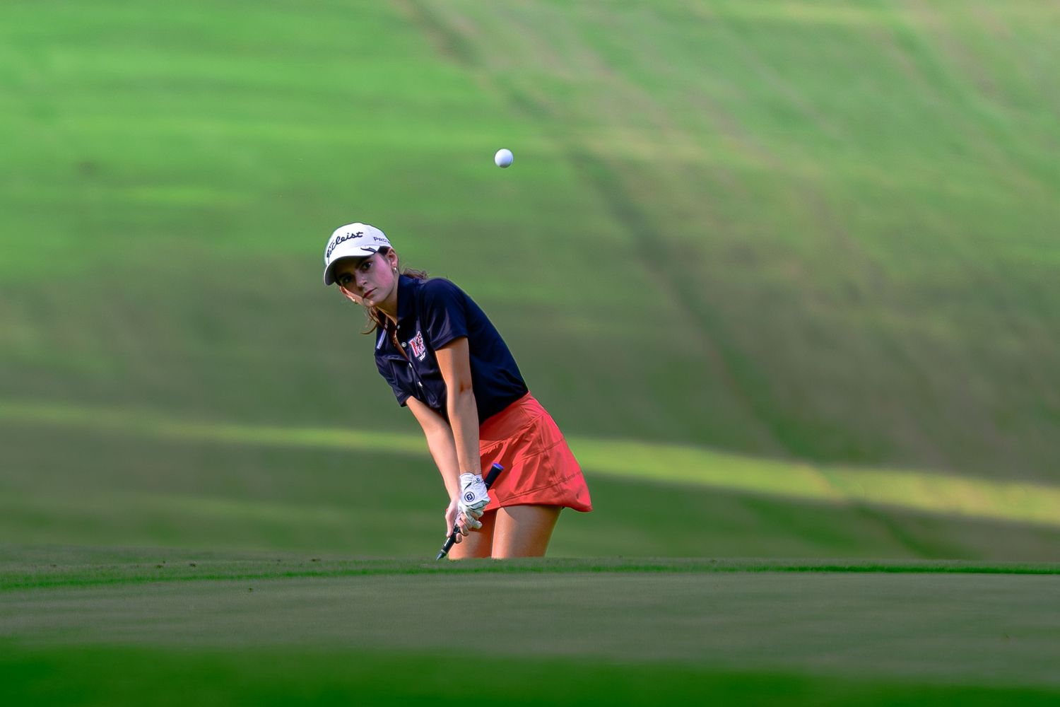 A female golfer in a dark polo and orange skirt follows through on a shot from the fairway.
