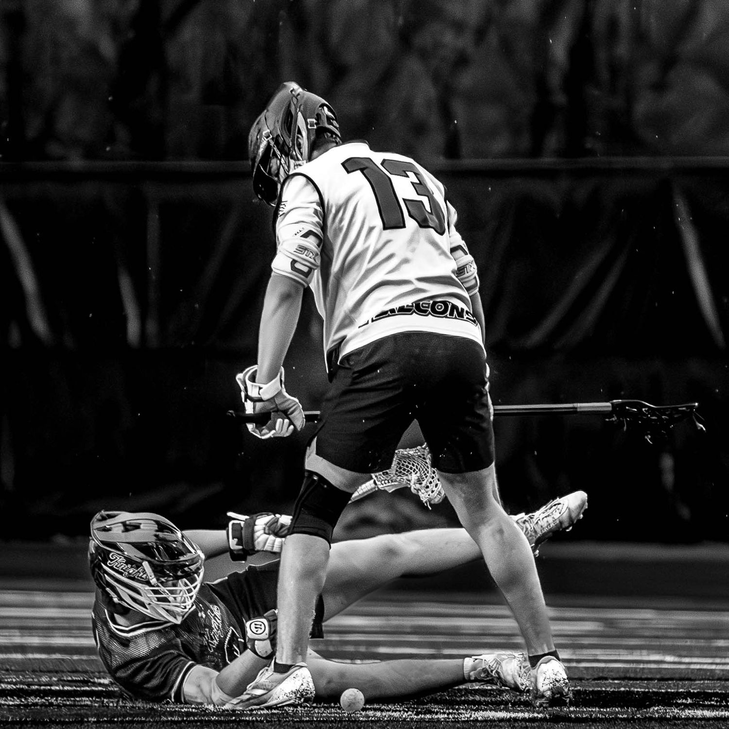 A black and white shot captures a high-intensity moment between two lacrosse players vying for possession on the turf.