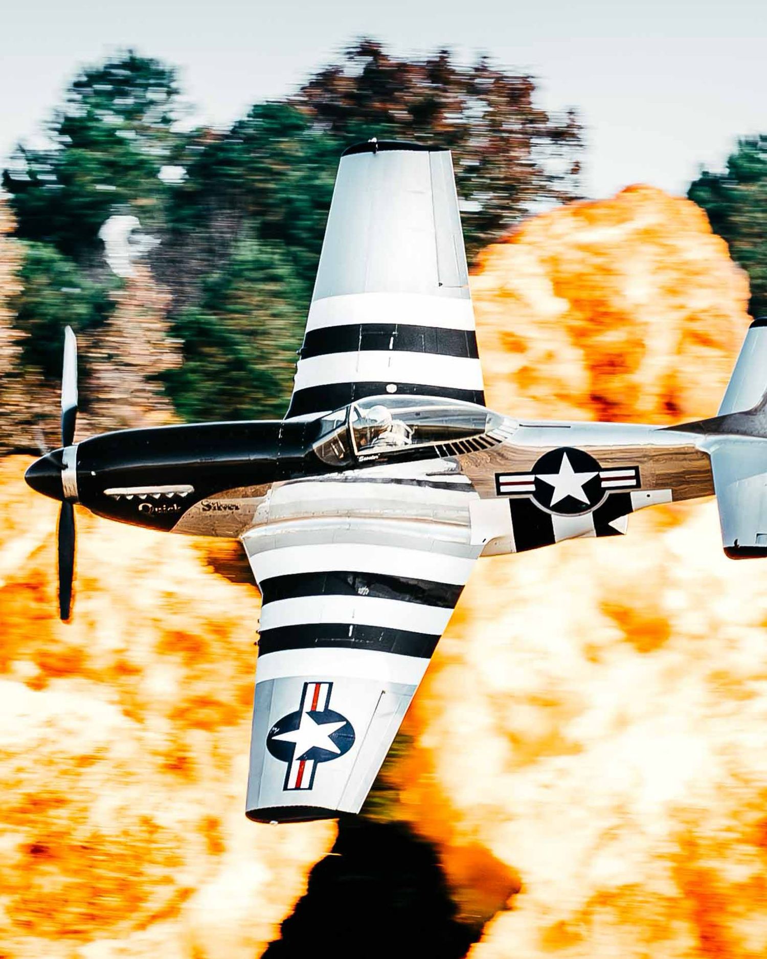 A vintage P-51 Mustang fighter plane flies past a massive wall of fire and explosions during an aerial stunt show.