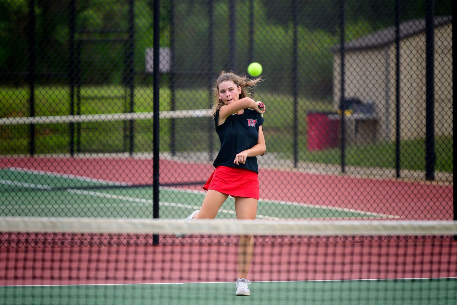 A female tennis player in a black top and red skirt prepares a powerful forehand return on an outdoor court.